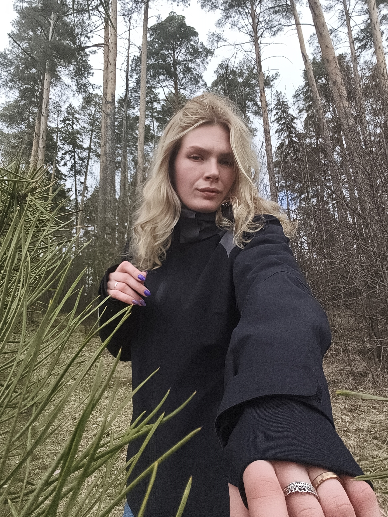 A fair-skinned woman with blonde, wavy hair stands in a wooded area. She wears a black jacket and has purple nail polish on her left hand. Several rings adorn the fingers of her outstretched right hand. Tall trees form the backdrop, with some dense green foliage visible on the left. The ground appears to be covered in dry grass or leaves. The picture's context indicates this is Izhevsk, Russia.