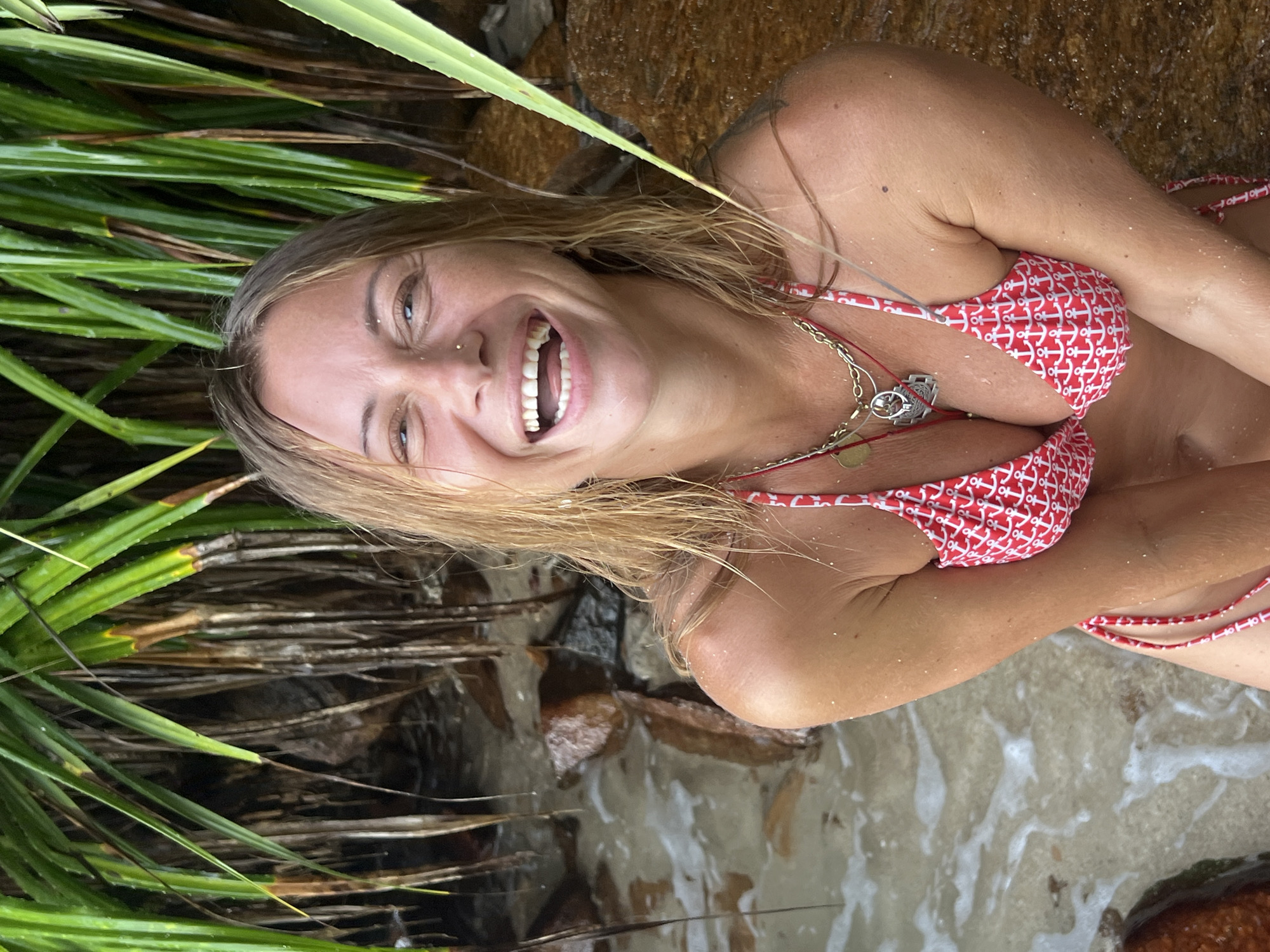 A young woman with blonde hair is lying on the sand with water lapping around her. She is wearing a red and white patterned bikini with an anchor design. Her eyes are closed, and she has a wide smile on her face, suggesting happiness or enjoyment. Her skin is lightly dusted with sand. She is adorned with several necklaces. In the background, lush green tropical foliage and rocks are visible, and the water appears clear and shallow. The scene suggests a sunny day in a tropical beach setting, likely Goviyana, Sri Lanka, based on the location context. The overall mood is relaxed and joyful.