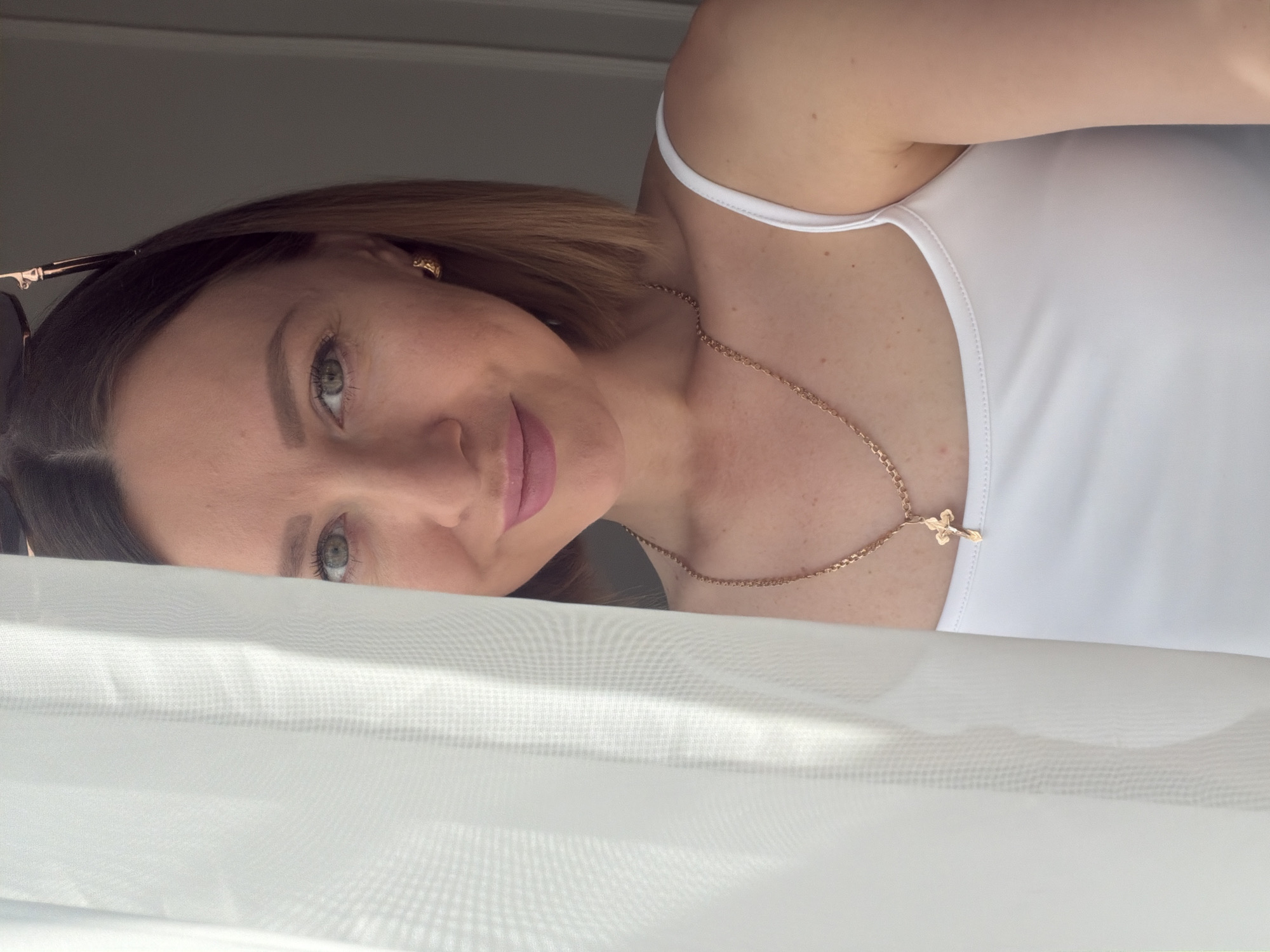 A woman with light skin, brown hair, and green eyes is partially visible, looking at the camera with a slight smile. She is wearing a white top and a gold necklace with a cross pendant. Sunglasses are placed on her head. A white, semi-transparent fabric is draped in the foreground. The location is not identifiable as Rostov-on-Don, Russia from the visual information provided.