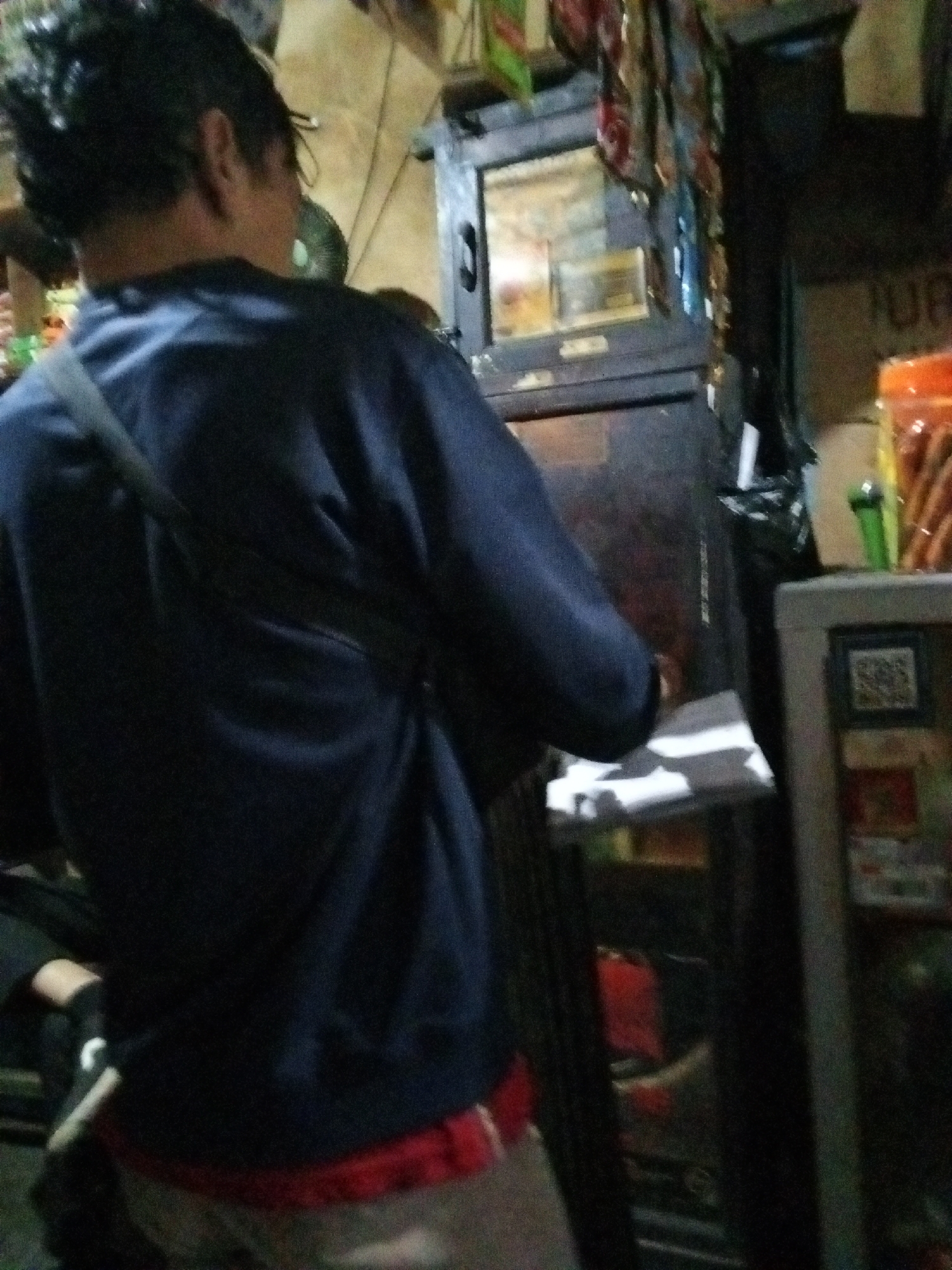 The image depicts a dimly lit interior scene, likely a small shop or warung in Bandung City, Indonesia. The primary subject is a person seen from behind, wearing a dark blue long-sleeved shirt over a red undergarment and light-colored pants. The person has dark, spiky hair and a strap from a bag crosses their chest. They are standing in front of a dark wooden cabinet or display case that has a glass-fronted section revealing some items inside. To the right, a small appliance with a QR code displayed on it is visible. Various goods, including what appear to be snacks and drinks in plastic packaging, are arranged on shelves and surfaces in the background. The overall atmosphere is cluttered and suggests a busy retail environment.