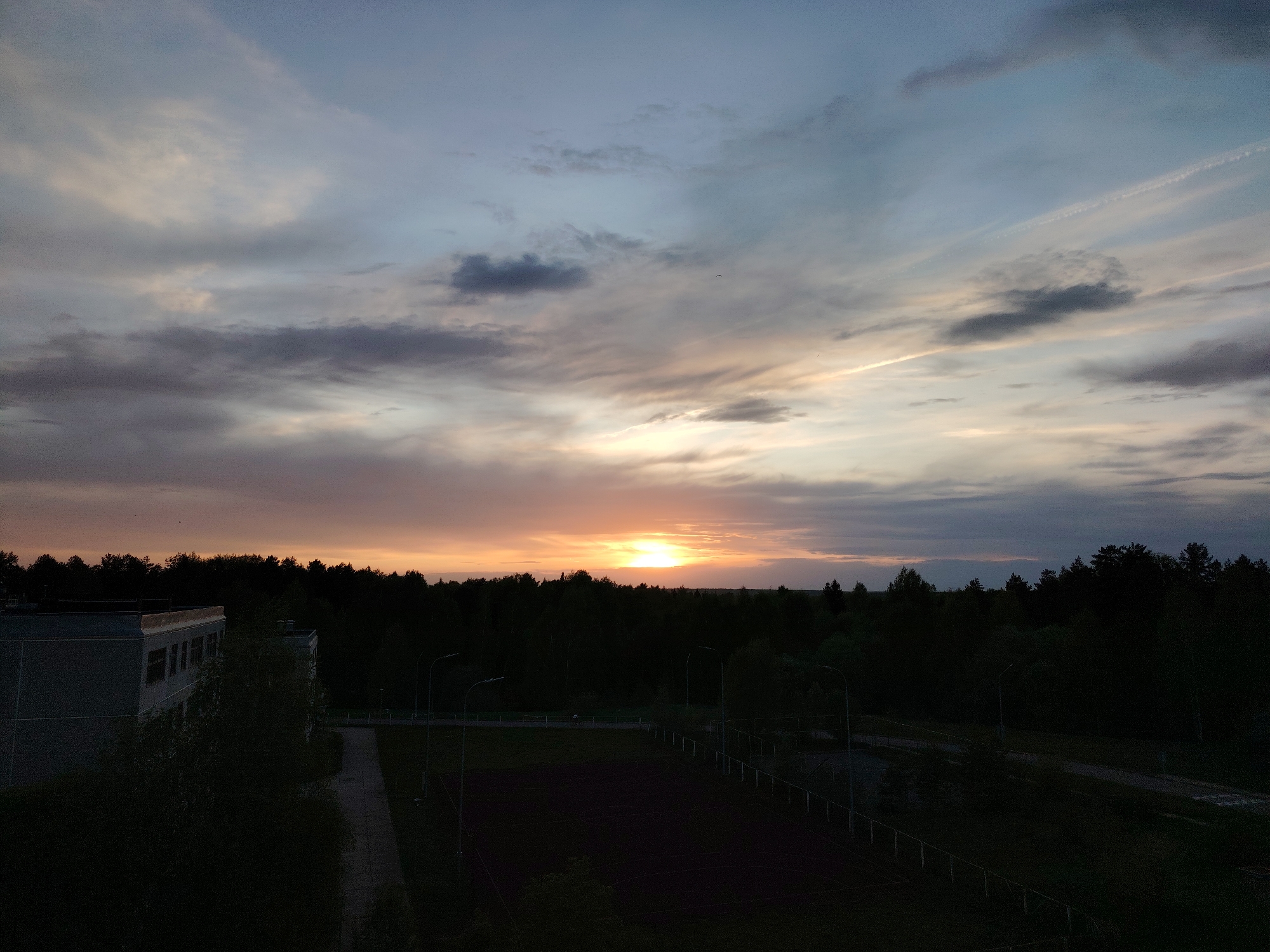 The image captures a sunset over Марковский, Russia. The sky is filled with clouds, ranging from dark grey to light blue, illuminated by the setting sun creating a gradient of orange and yellow hues. Below the horizon, a dense forest line separates the sky from the foreground. A building is visible on the left side of the image, appearing silhouetted due to the low light conditions. A sports field or a similar open area with fence is positioned in front of the forest. Several street lights are visible, contributing to the minimal illumination of the scene. The overall environment appears calm and serene.