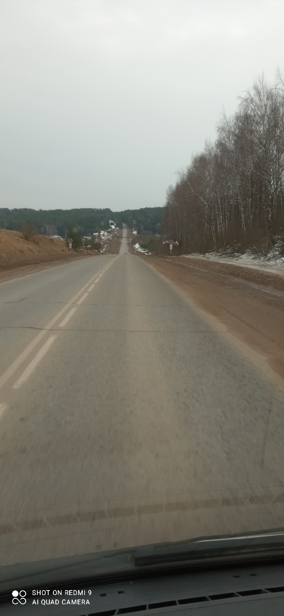 The image depicts a road in Ольховка, Russia, as seen from inside a vehicle. The road is asphalt, with a double white line dividing the lanes. The road appears to be heading uphill toward a small village. Patches of brown grass and trees without leaves are visible on either side of the road. The sky is overcast. A traffic sign is located on the side of the road. The dashboard of the vehicle is visible at the bottom of the frame.