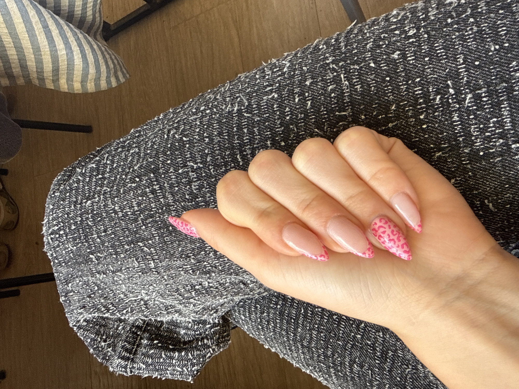 A close-up, eye-level shot shows a left hand with almond-shaped nails with a French manicure and pink leopard print tips, resting on a person's lap. The person is wearing dark, textured trousers. In the background, a table with a blue and white striped cloth is visible. The scene is indoors, with wooden flooring. The location is not specified within the city of Sofia, Bulgaria.