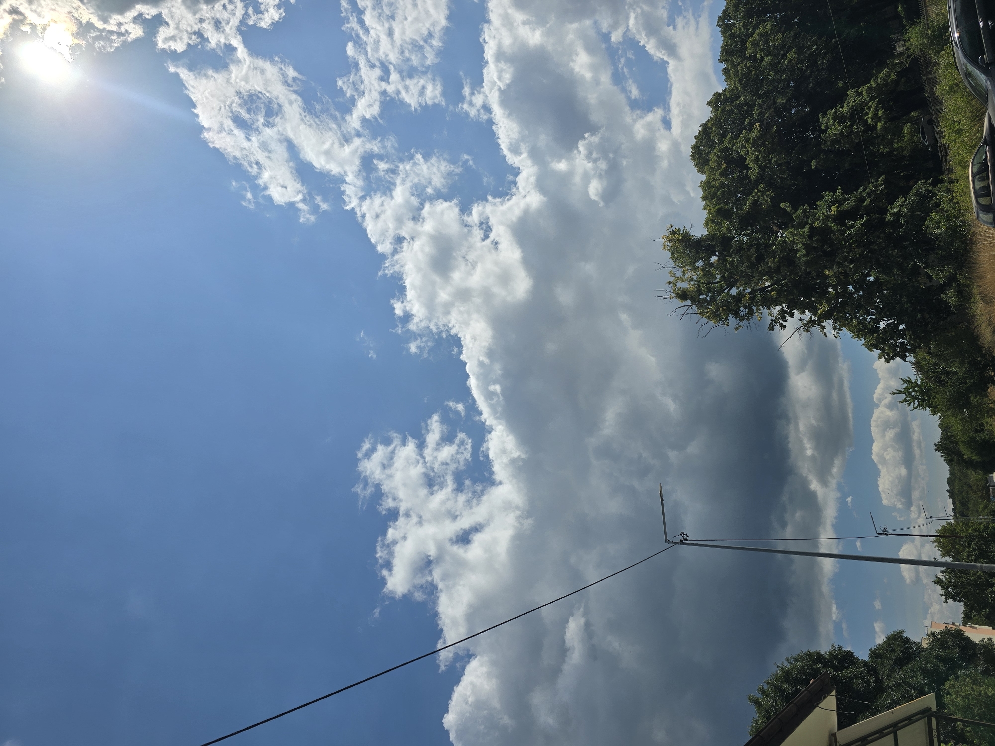 The image depicts a bright, partly cloudy sky over a semi-rural area, possibly on the outskirts of Malko Tarnovo, Bulgaria. A power line stretches diagonally across the upper left of the frame. In the mid-ground, a tall street lamp stands prominently, with another shorter one visible further down the road. Dense green trees line the right side of the image, and to the left, a portion of a building with a light-colored roof is visible. A few cars are parked or driving along a road in the lower portion of the scene. The sun is visible in the upper right corner, partially obscured by clouds, casting bright light and lens flare. The overall impression is a daytime scene, likely in the summer, characterized by a mix of natural and man-made elements.