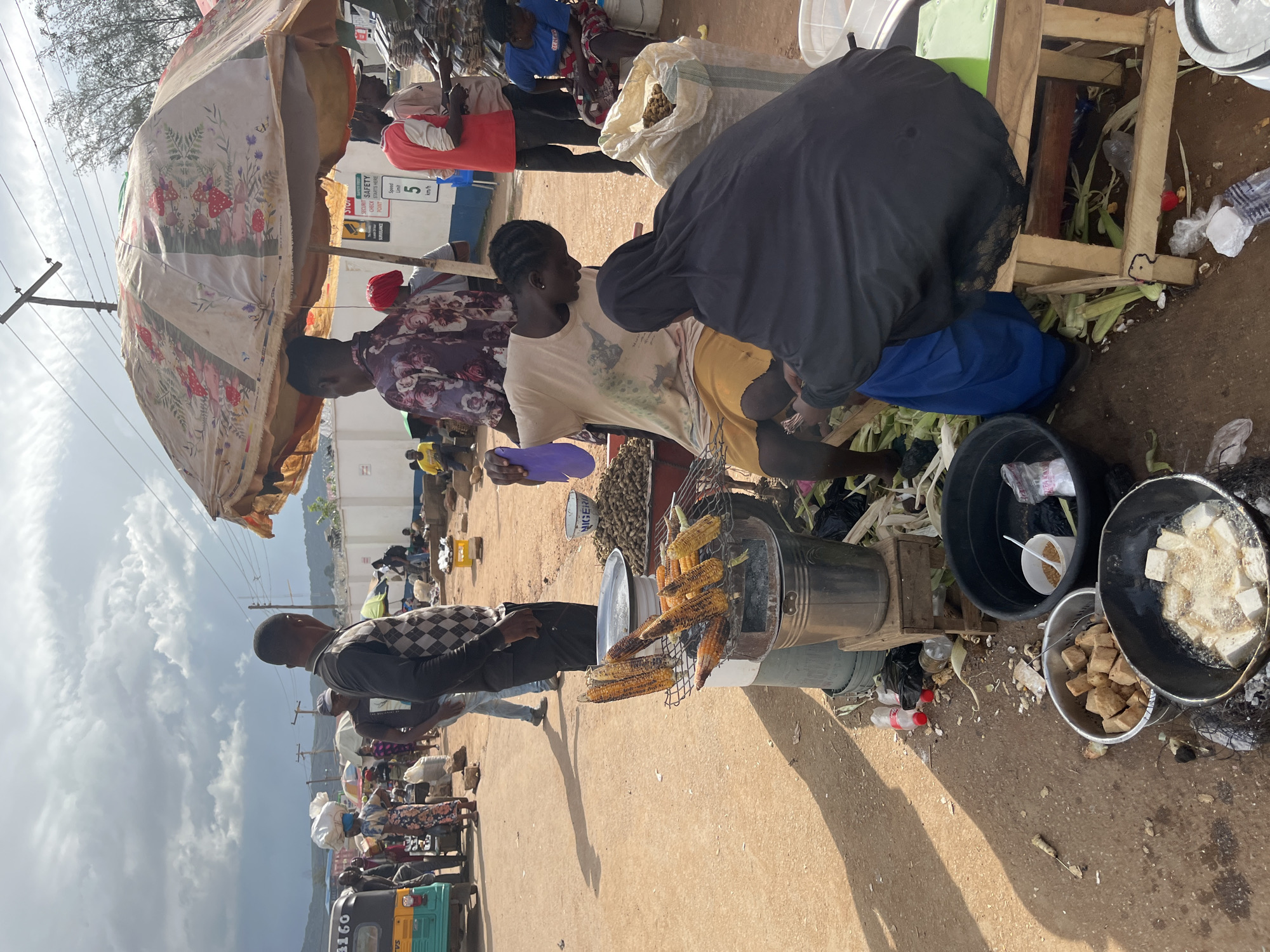 This is a photo of an outdoor market scene in Rafin Par, Nigeria. In the foreground, a man in a checkered shirt is standing, looking to the right. To his left, a young woman wearing a patterned shirt is standing, holding a purple plate. A woman in a black hijab and a blue skirt is leaning over a wooden structure, appearing to interact with the woman in the patterned shirt. In front of them, ears of corn are roasting on a grill over a metal bucket. Nearby, a black bucket and a metal bowl contain what appears to be fried food. Other people are visible in the background, along with stalls and various market goods. The scene is set on a dusty ground under a partly cloudy sky, suggesting it's daytime. The overall atmosphere appears to be that of a busy marketplace. A yellow and green tricycle is parked in the lower left corner.