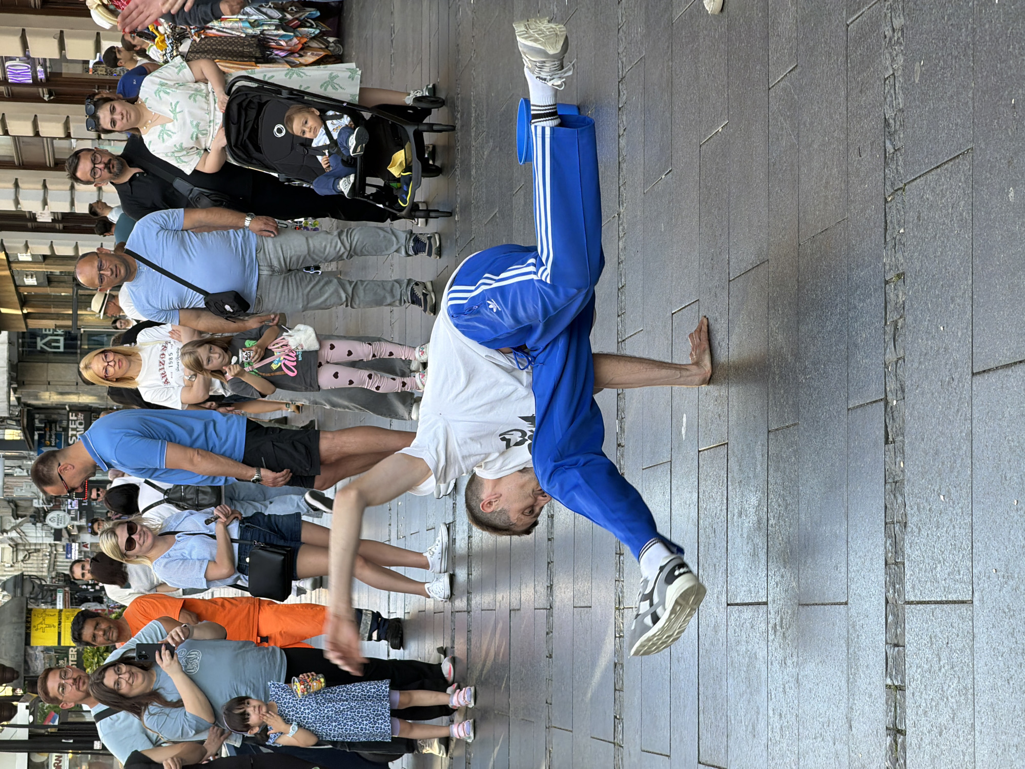 A man in a white T-shirt and blue Adidas tracksuit is performing a handstand on a gray-tiled sidewalk in Belgrade, Serbia. He is barefoot on his right hand. He is surrounded by onlookers, including a family with a baby in a stroller, individuals in casual attire, and people holding phones and cameras. The sidewalk is made of large rectangular tiles, with a narrow strip of smaller cobblestone tiles running parallel to the main surface. In the background are buildings with shopfronts.
