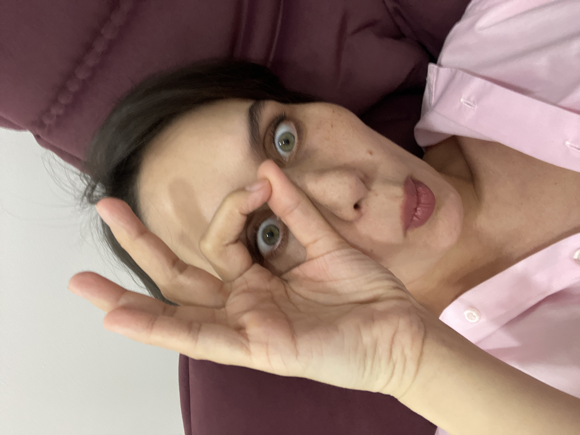 A woman is seen lying down with her head tilted towards the camera, making an "OK" sign with her hand over her eye. She has dark hair and fair skin, and is wearing a pink button-down shirt. Her eyes are wide, and she has a somewhat surprised expression. She is lying on a maroon-colored cushion or couch. The background is a plain white wall. There is no information in the image to indicate a location within the city of Izhevsk, Russia.