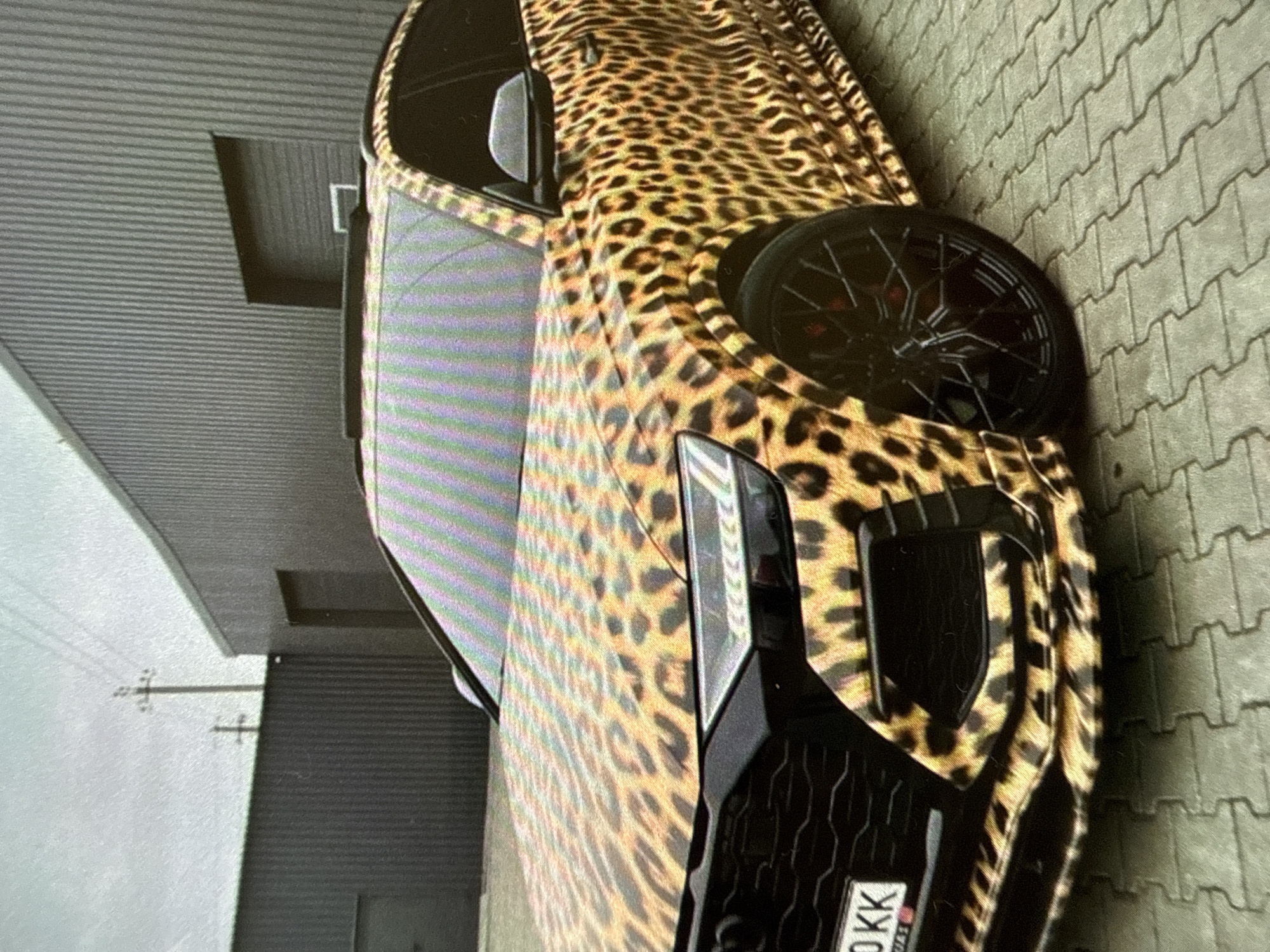 Here is a description of the image:
A car with a leopard print wrap is parked outdoors in an urban area. The car is viewed from the front and side, showcasing the wrap's pattern. The vehicle has black wheels with red brake calipers visible behind the spokes. In the background, there's a building with gray siding and rectangular windows. The ground is paved with interlocking gray bricks. Overhead power lines are visible in the distance, further placing the scene in a populated area.