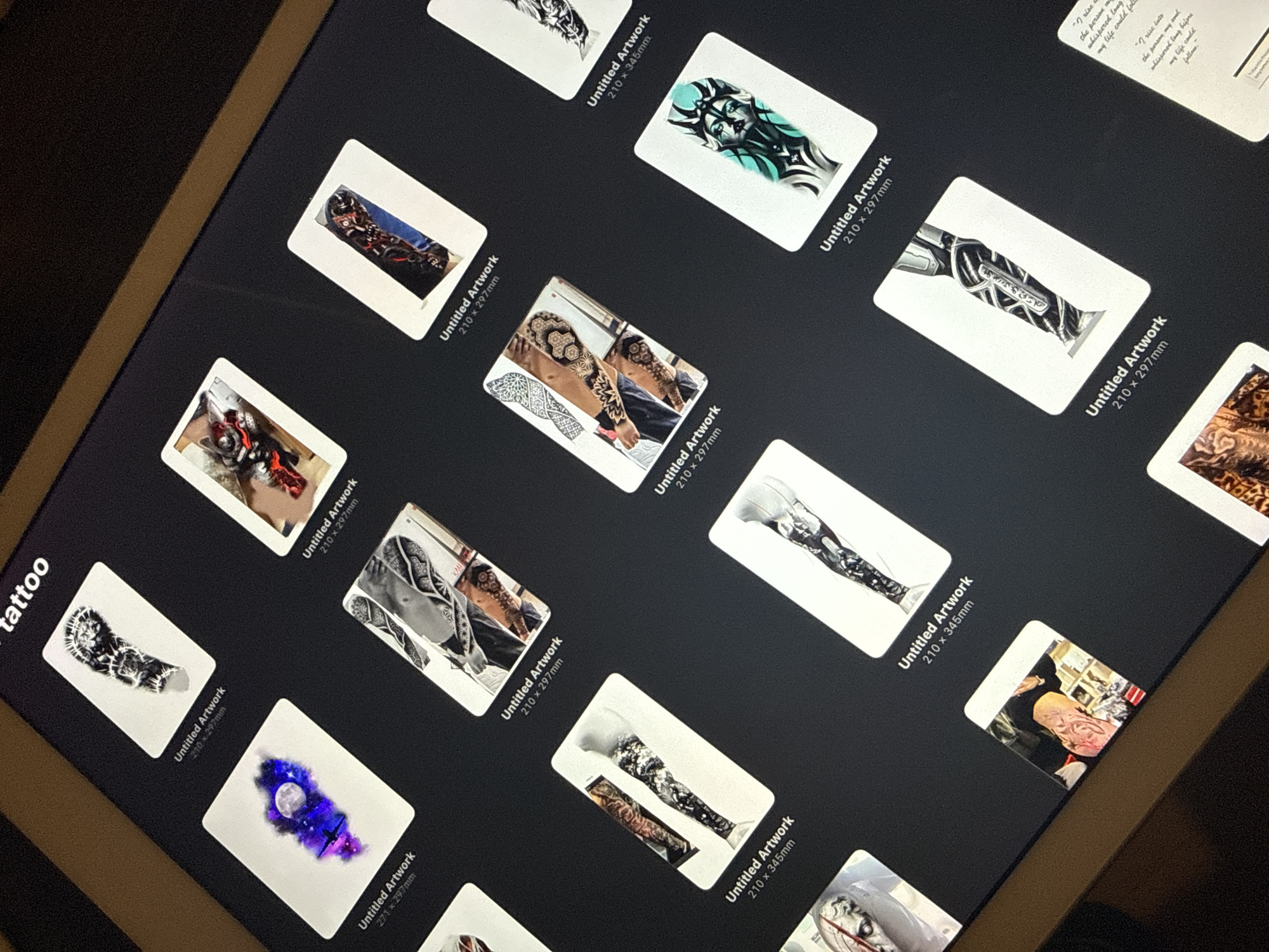The image shows a digital screen, likely of a tablet, displaying a collection of digital artwork thumbnails. The artworks are presented as white rectangles against a dark background, each labeled "Untitled Artwork" with dimensions such as "210 x 297mm" or "210 x 345mm". The artwork itself consists of various images and designs. One thumbnail contains a complex geometric pattern on an arm, while others showcase images of arms, legs, or body parts adorned with intricate designs. Other thumbnails display images that include a moon and other celestial objects, mechanical parts, and a face with teal markings. The word "tattoo" is visible on the left side of the screen. The location of this scene is not determined.