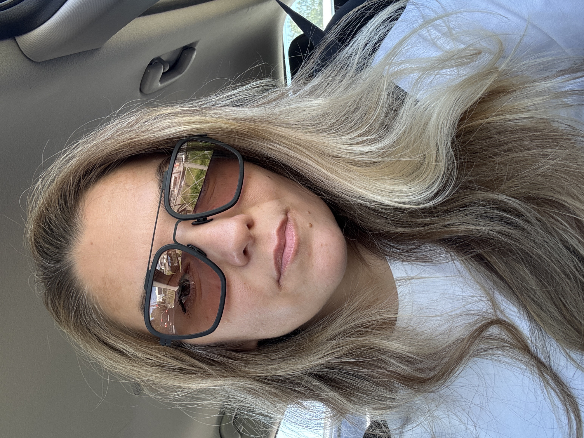 A close-up, selfie-style shot shows a woman wearing dark-rimmed sunglasses and a white t-shirt. She has blonde hair with lighter streaks, which is styled in loose waves and falls around her shoulders. Her skin appears fair with a scattering of freckles on her forehead and cheeks, as well as a small mole on her chin. The sunglasses reflect a scene of what appears to be a street with cars and buildings. The background suggests the interior of a car, with a grey headliner and a black car handle visible. The lighting indicates it is daytime, likely outdoors. The image does not provide any specific details to place it within the city of Saint Petersburg, Russia.
