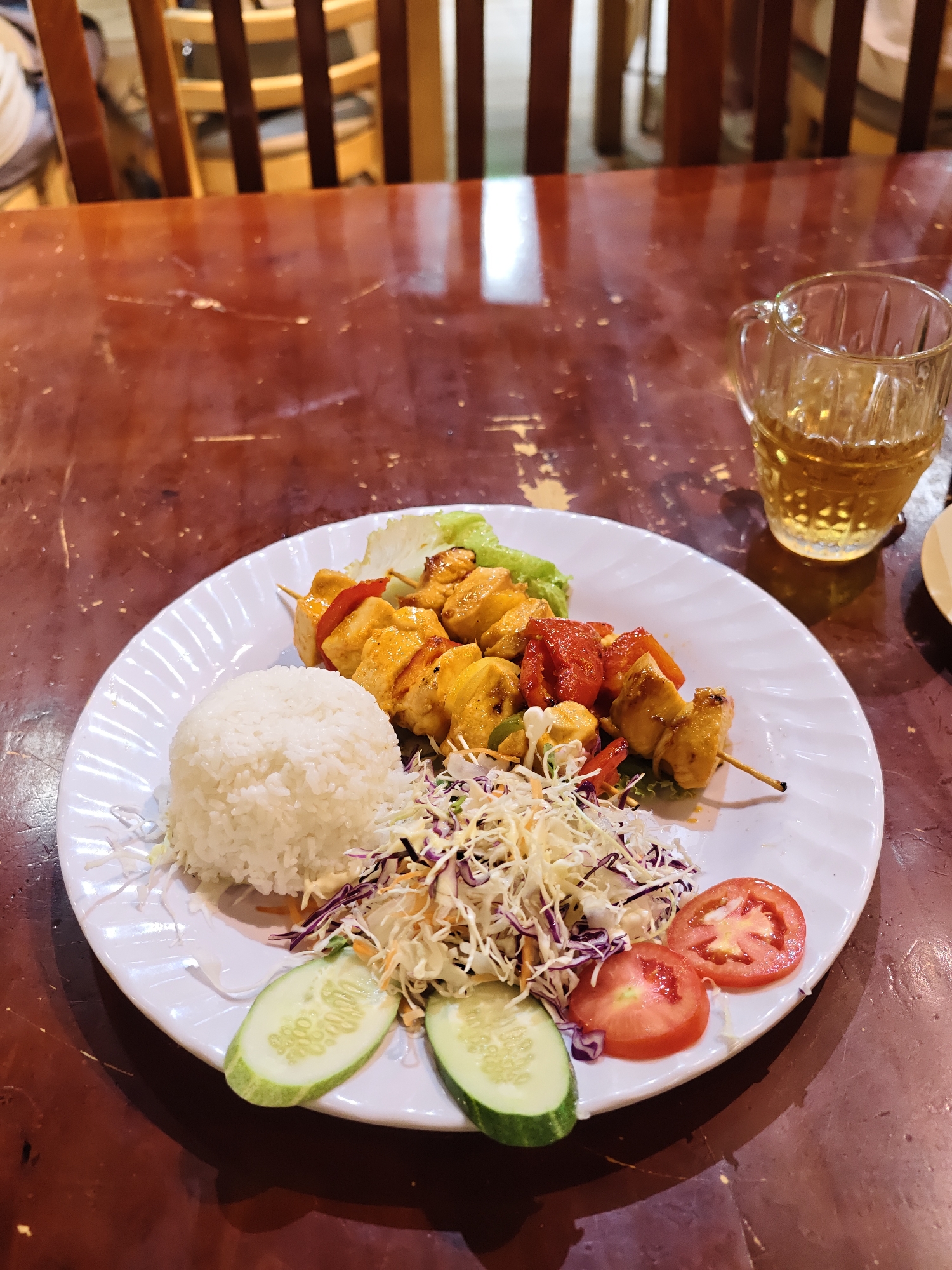 A close-up, eye-level shot shows a white plate of food on a dark wooden table. The plate contains a serving of white rice, a small salad with shredded cabbage and carrots, and two skewers of grilled chicken and vegetables. The skewers are topped with bell peppers and other ingredients, and appear to be glazed. The salad is garnished with sliced cucumbers and tomatoes. To the right of the plate, a clear glass mug filled with a light-colored beverage, possibly iced tea, is visible. The background shows wooden chair backs, suggesting a restaurant setting. The lighting appears to be artificial, with warm tones reflecting off the wooden table, indicating an indoor environment. The food looks fresh and appetizing. There is no visible text in the image.