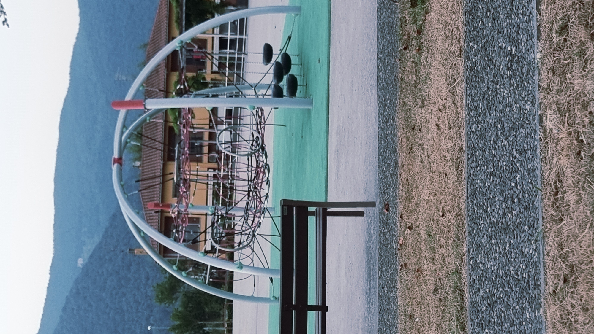 The image depicts a playground located in La Canya, Spain. In the foreground, a dark brown park bench is positioned on a grey paved surface. Beyond the bench, a vibrant green rubberized playground surface is visible, featuring a modern climbing structure with a geodesic dome frame. This structure is made of white metal poles and interconnected ropes, with several black tire swings hanging from it. In the background, a light yellow building with a terracotta roof is situated amongst trees. The scene is set against a backdrop of lush green mountains under a bright, hazy sky. The overall environment suggests an outdoor public space, likely a park or recreational area.
