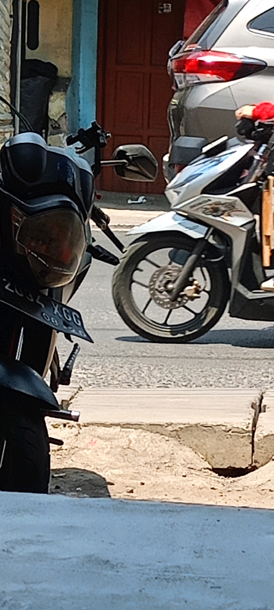 A dark-colored motorcycle is parked on the right side of the frame, its front wheel and handlebars partially visible. To the right of this motorcycle is a silver SUV. In the background, a reddish-brown door is visible on a building with blue accents. The foreground shows a cracked concrete curb. A license plate on the dark motorcycle reads "2034 XGG".