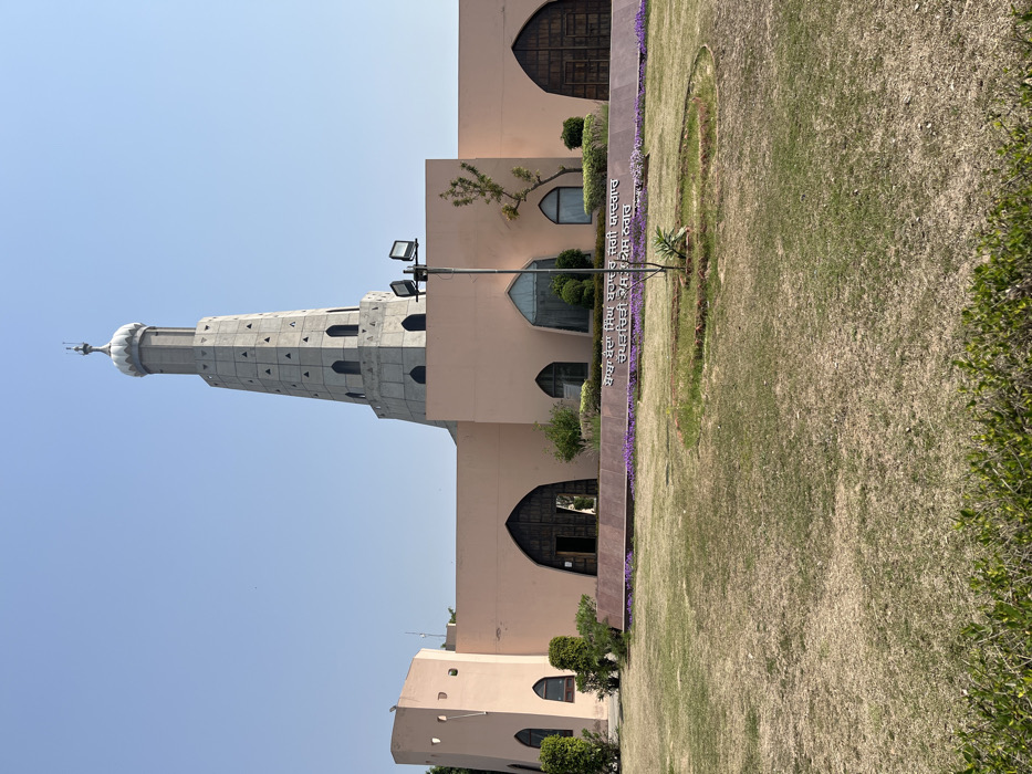 The image shows a large, pale pink, rectangular building with a tall, cylindrical tower in the center. The tower is adorned with a white dome. The building has multiple arched windows with a large arched doorway at the center. The building is surrounded by green grass and a few purple flower patches. In front of the building, there is a sign with text in an unknown language, likely Punjabi, and two light poles with dual lights. The scene is taken in Sahibzada Ajit Singh Nagar, India, at the coordinates 30.703 latitude and 76.668 longitude. The sky is clear and blue.