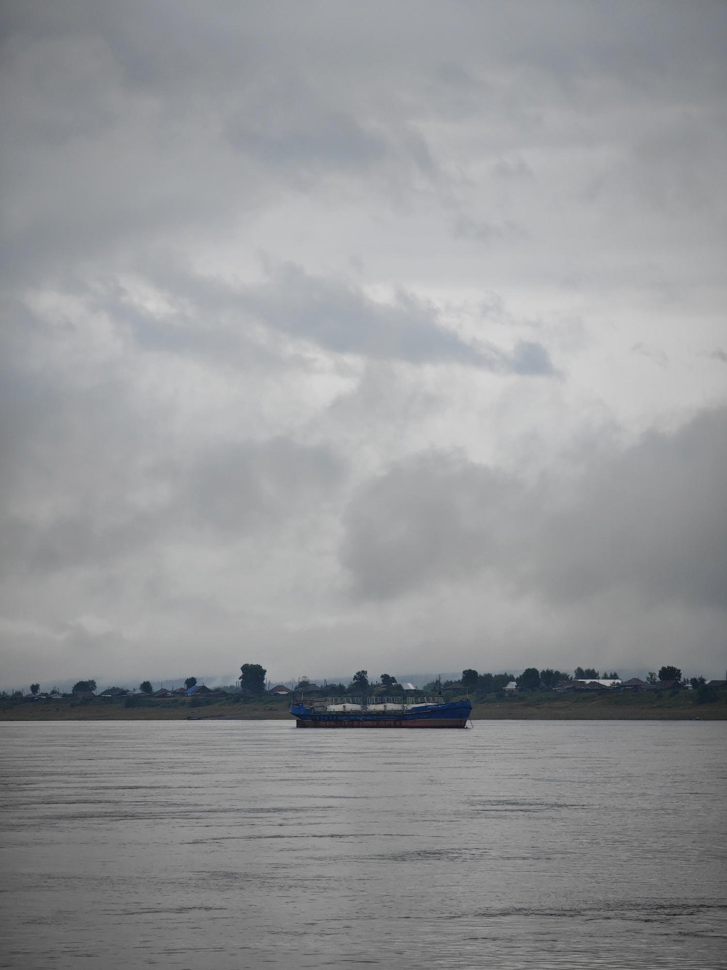 A barge is moving on a wide, calm river under a heavily overcast sky. The sky is filled with gray, textured clouds, with some lighter patches suggesting a break in the cloud cover. Along the distant bank, a line of trees and a few scattered buildings are visible, indicative of a settlement. The water is a muted gray and shows faint ripples. The barge is blue and red, with white railings, and appears to be laden with cargo. The overall atmosphere is somber and quiet. The location is the Angara River in Lesosibirsk, Russia.