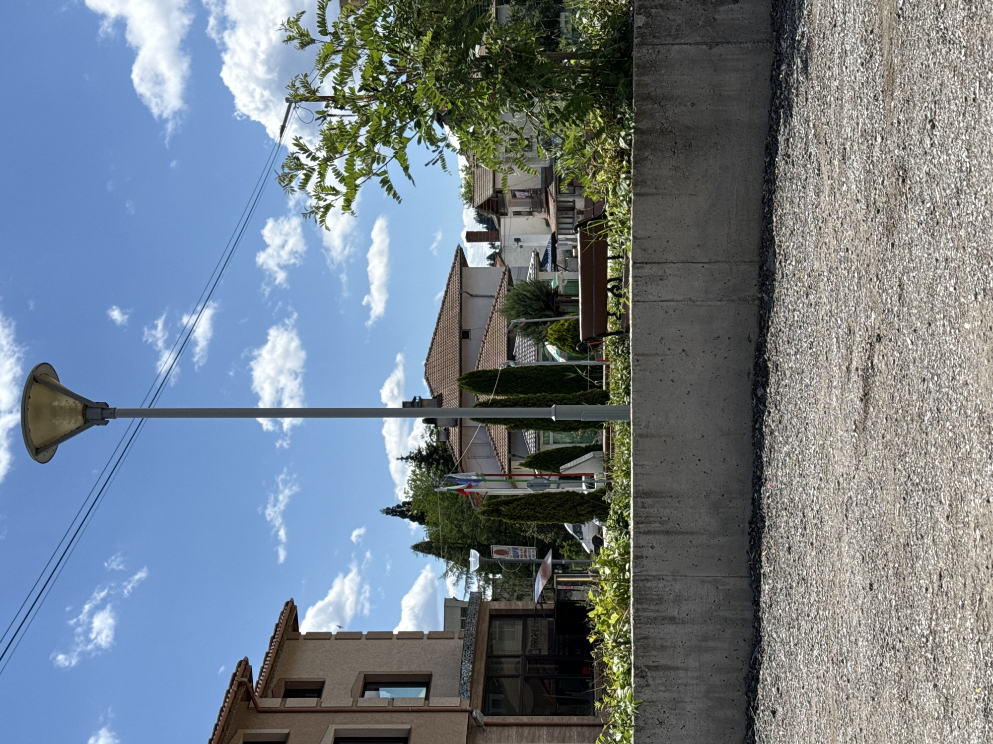 The image captures a street view in the city of Barutin, Bulgaria, under a bright, cloudy sky. In the foreground, a gravel surface leads up to a concrete retaining wall. Behind the wall, a lush green garden with various plants is visible, including a prominent tree on the right. A simple wooden bench sits in the garden, facing away from the viewer.
Several buildings are present in the midground. On the left, a multi-story building with a light-colored facade and dark-rimmed windows is partially visible. In the center, a street lamp with a distinctive yellow shade stands tall. Next to it, flags are flying from poles. Various other buildings with tiled roofs, some appearing residential, are spread across the background. A white van is parked on the street. The overall environment suggests a typical urban or semi-urban setting with a mix of residential and commercial structures.