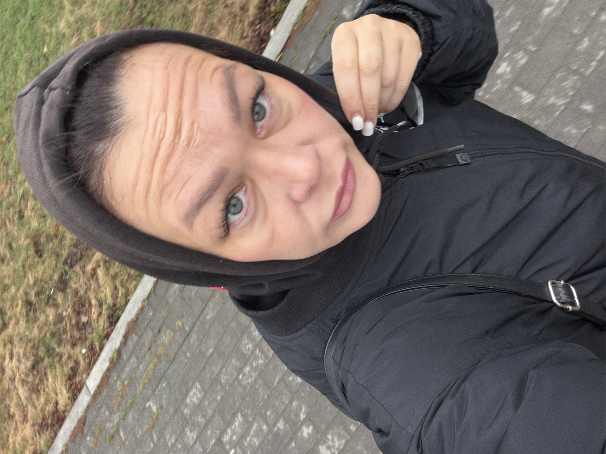This is a close-up, tilted selfie of a woman taken outdoors, likely in Chaikovsky, Russia. She appears to be in her 20s or 30s, with fair skin, prominent light-colored eyes (greenish-blue), and dark hair visible beneath a dark, hooded jacket. Her expression is subtle, perhaps pensive or slightly thoughtful, and her forehead shows some natural lines. She is dressed in a black hooded puffer jacket, and the strap of a dark bag with a silver buckle is visible over her right shoulder. Her right hand is raised near her face, with white or light-colored manicured nails.
The background consists of a paved walkway, possibly made of grey pavers, beside a patch of dry, light-brown grass and dormant vegetation. The natural, diffused lighting suggests an overcast day, and her warm attire points to cooler temperatures, such as those found in late autumn, winter, or early spring. No visible text is present in the image.