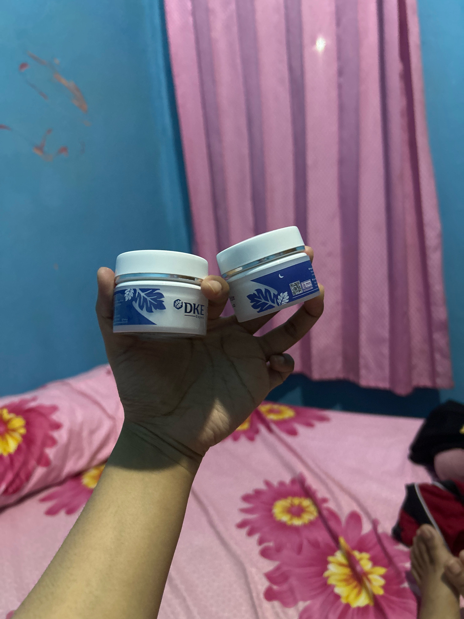 A person's hand holds two small white jars with blue and white labels. The jar in the foreground has the text "DKE" and "Expert" visible on its label, along with a blue leaf design. The jar behind it has a similar design and a crescent moon symbol. The background features a teal-colored wall with some orange splatters and pink curtains. Below the hand, a bed with pink floral-patterned bedding is visible, and part of a person's leg and foot can be seen. This scene appears to be in a bedroom, possibly in the city of Cihaurbeuti, Indonesia.