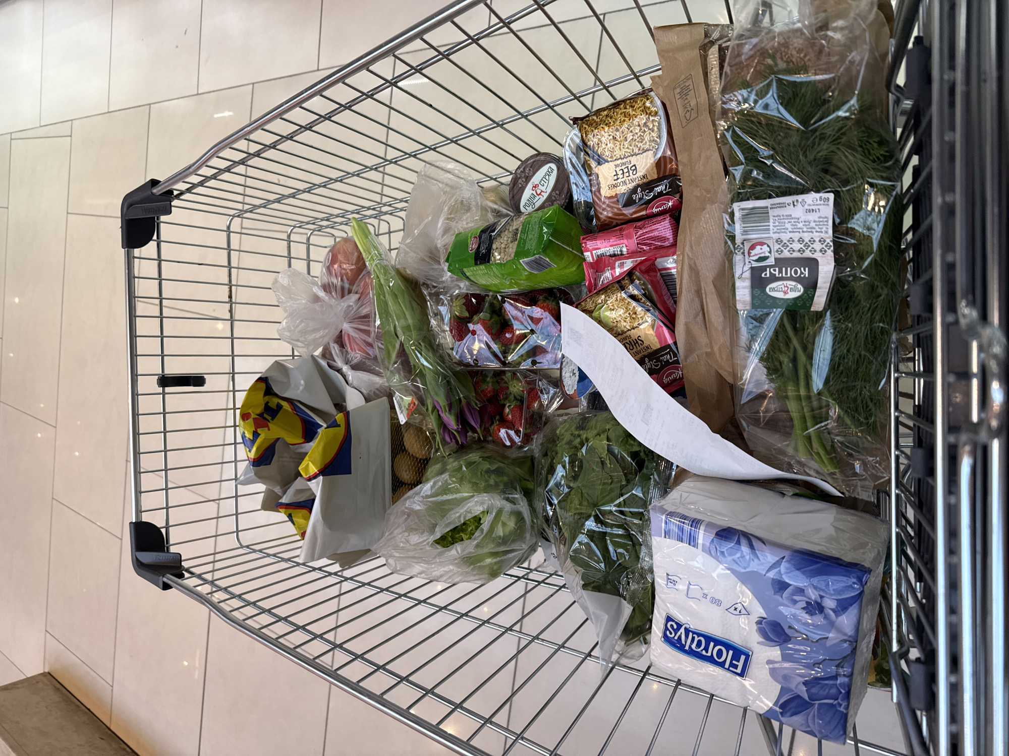 A shopping cart is filled with various grocery items, including packaged dill ("КОПЪР"), a box of "Floralys" tissues, strawberries, lettuce, kiwis, red onions, instant noodles (beef and shrimp flavor from "Kania" brand), lentils, and a round cheese. The background features a light-colored tiled floor, suggesting the setting is within a grocery store. Given the Cyrillic text on the dill packaging ("РОДНА СТРЯХА" and "КОПЪР"), and the common availability of brands like "Floralys" and "Kania," a plausible location for this scene is Sofia, Bulgaria.