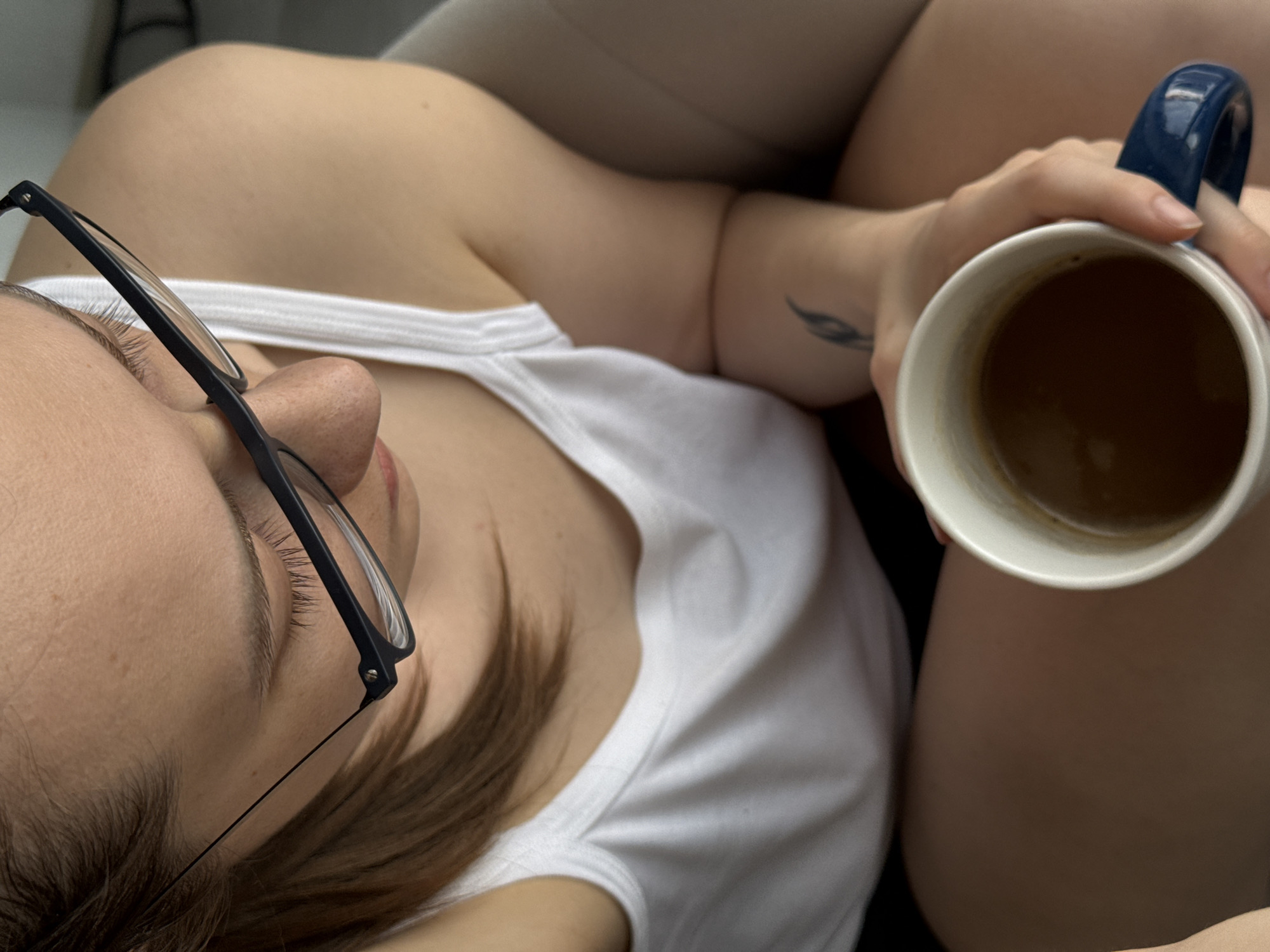 The image displays a female subject in an indoor setting in the city of Chaikovsky, Russia, captured from a high-angle perspective looking down. The subject is wearing a white tank top and dark-colored shorts or similar lower garment. She has light brown hair and is wearing dark-framed eyeglasses positioned on her nose. Her gaze is directed downwards. A dark, stylized tattoo resembling a feather or leaf is visible on her left forearm. Her right hand holds a white ceramic mug with a dark blue handle, containing a dark brown liquid, consistent with coffee. The immediate background consists of indistinct, light-toned surfaces, suggesting a domestic environment.