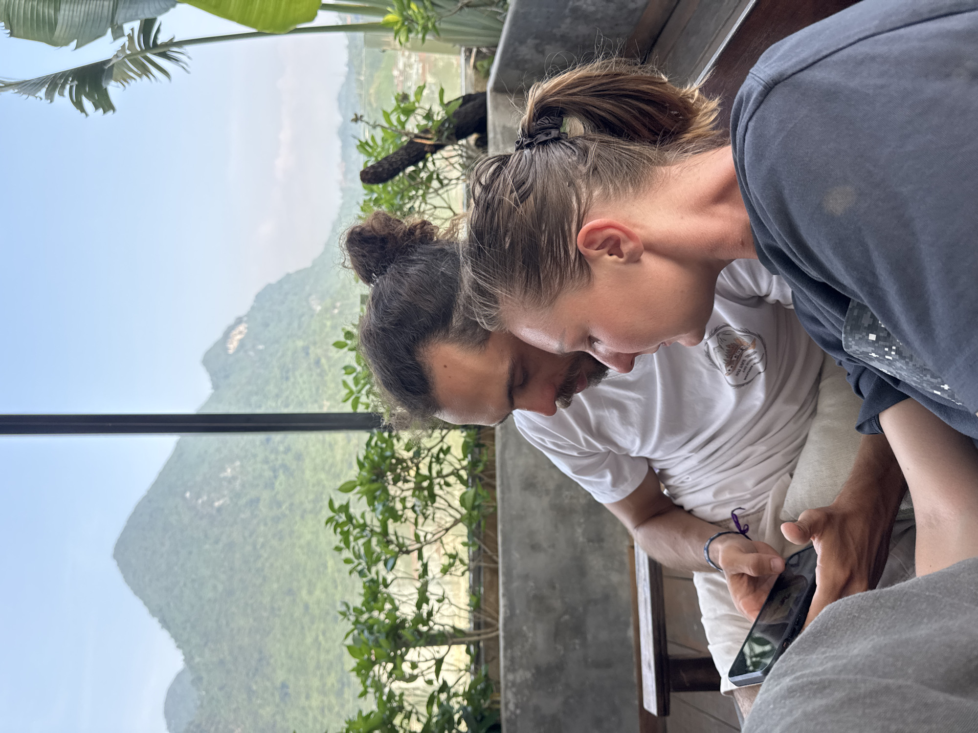 The image, oriented correctly, depicts two individuals intently focused on a smartphone in a scenic indoor setting. In the foreground, a female with her hair tied in a ponytail wears a dark long-sleeved top and holds a black-cased smartphone, her gaze directed downwards at the screen. Behind her and slightly to the left, a male with dark hair in a bun and a beard also leans in, looking at the phone. He wears a white t-shirt with a faint, indecipherable logo and a purple bracelet on his left wrist.
The two appear to be sharing or viewing content on the device. They are seated, likely on a bench, next to a modern concrete ledge or wall that separates them from a stunning panoramic view. Outside, through large windows or an open balcony, lush green mountains dominate the landscape. The sky is bright and clear, suggesting daytime, with a soft haze over the distant peaks. Green foliage, including large palm-like leaves, is visible close to the viewing area and further out on the mountain slopes, indicating a verdant environment typical of Hà Giang 1 Ward, Vietnam. The scene conveys a moment of shared concentration amidst a beautiful natural backdrop. No specific text is clearly legible in the image.