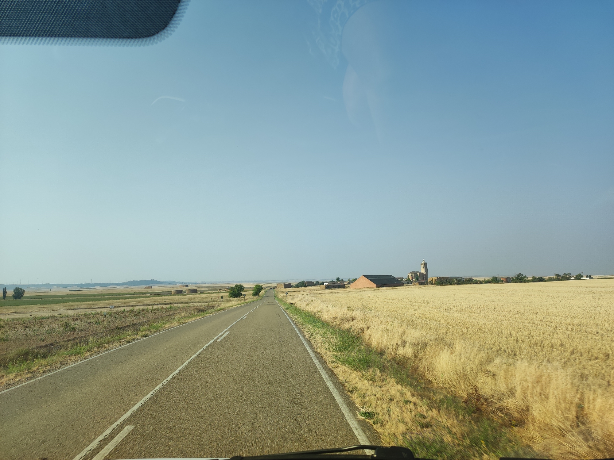The image, taken from inside a vehicle, shows a paved road stretching through a rural landscape. The road is flanked on the right by a field of dry, golden wheat or barley. On the left, the landscape consists of cultivated fields and some scattered trees. In the distance, on the right side of the road, a village is visible with buildings, including a church with a tower. Wind turbines are present on the horizon. The sky is clear and blue. The image appears to have been taken in the vicinity of Baquerín de Campos, Spain, judging by the characteristic agricultural landscape and architectural style of the village.