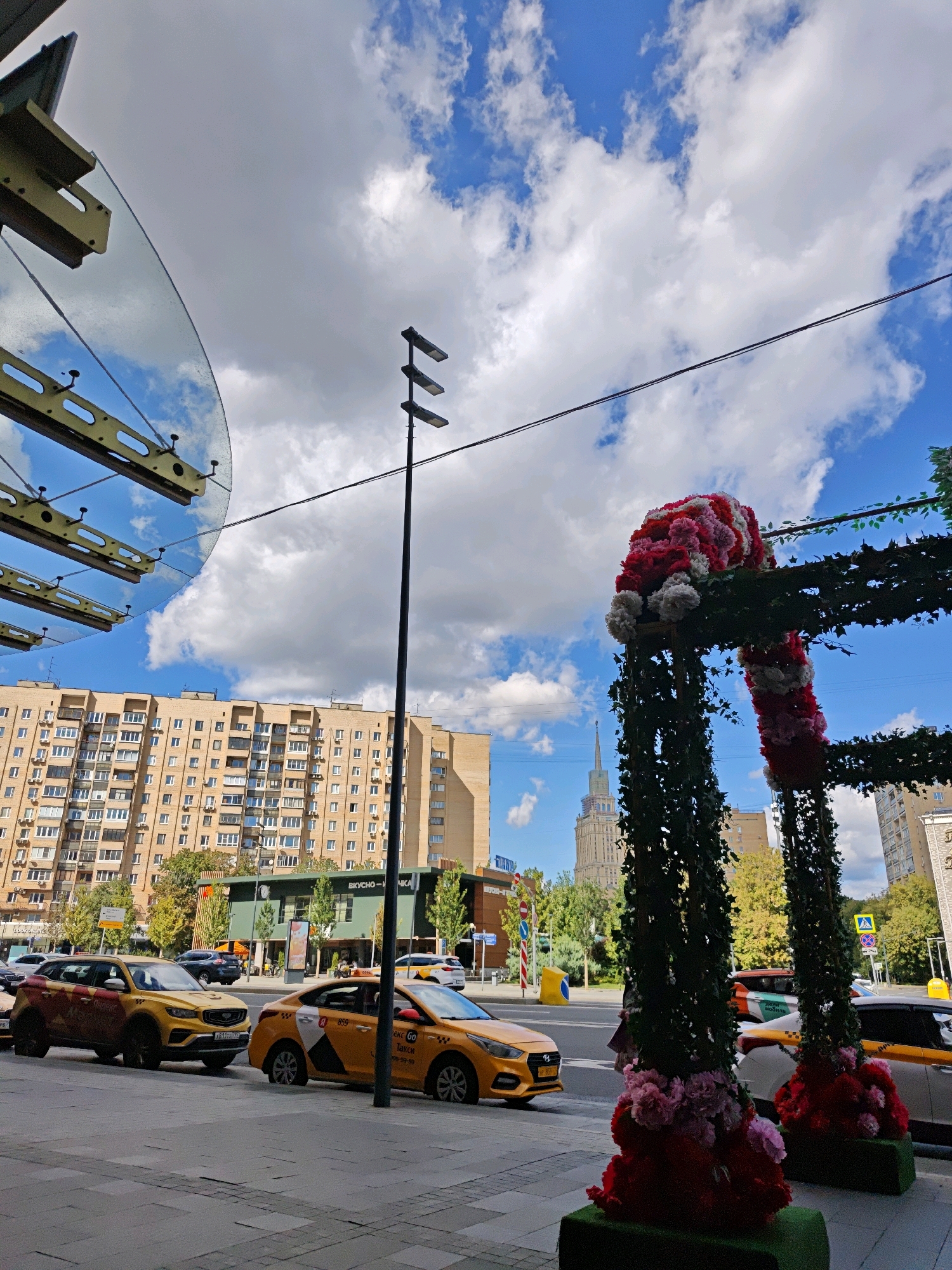 This is a medium-shot, eye-level, outdoor photograph taken on a sunny day in Moscow, Russia. The scene is a street with a sidewalk in the foreground. Two yellow taxis are parked on the street. The taxi on the right is a Yandex Go branded vehicle. Behind the taxis, there is a building with many windows and a sign in Russian. Further in the background, a tall, Stalinist-era skyscraper with a spire dominates the skyline. In the right side of the frame, a large decorative archway covered in ivy and flowers is present. The sky is blue with scattered white clouds. The overall atmosphere is urban and bustling, with elements of both modern and historical architecture.