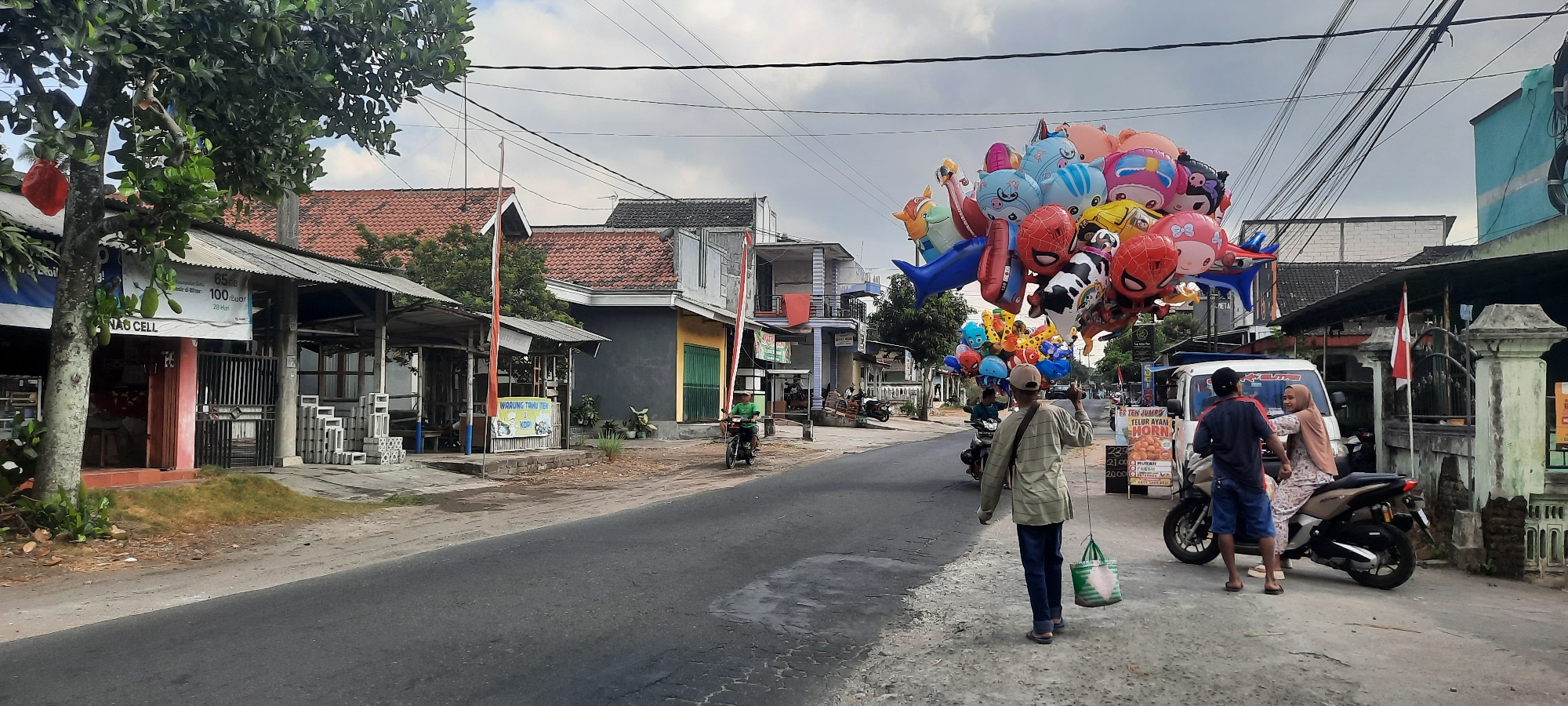 In Jatitengah, Indonesia, on an overcast day, a street vendor walks along the side of a paved road, carrying a large bouquet of colorful, character-shaped balloons. The balloons are in various designs, including Spider-Man, SpongeBob SquarePants, and animal shapes. He holds a string attached to the balloons, and a green and white reusable bag hangs from his left hand. He is wearing a striped long-sleeved shirt, dark pants, and sandals.
The street is lined with buildings, some with tiled roofs and others with flat roofs. On the left side of the road, a small shop with a red awning and a sign that reads "NAB CELL" is visible. Further down the street, a small structure with a thatched roof appears to be a food stall or warung. A person on a motorcycle rides past this stall.
On the right side of the road, a white van is parked, with a man and a woman standing next to it. The woman is wearing a hijab and a patterned dress, and is seated on a scooter. The man is wearing a dark t-shirt and blue shorts. A sign behind the van advertises "TEN JUMBO TELUR AYAM". Several motorcycles are parked along the street. The overall environment suggests a typical street scene in a residential or commercial area of a town in Indonesia.