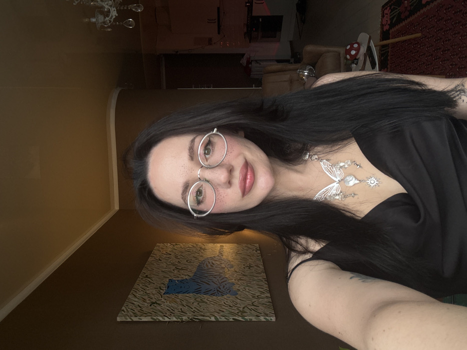 The image depicts an indoor scene within the city of Batumi, Georgia, focusing on a single female subject. The subject is positioned centrally, facing forward, and appears to be taking a self-portrait. She has long, dark hair, fair skin with visible freckles across her nose and cheeks, and wears oval-shaped, silver-rimmed glasses. Her lips are painted with a light pink shade, and she exhibits a subtle smile. She is dressed in a black satin top or dress with a cowl neckline and wears a prominent silver-toned necklace featuring a large butterfly design, multiple beads, teardrop-shaped pendants, and a sun motif. A portion of a tattoo is visible on her left upper arm.
The background features warm brown walls and a light-colored ceiling with white crown molding. To the subject's left, a framed artwork on the wall displays a blue tiger-like figure amidst green and yellow vertical strokes, suggesting grass. To the subject's right, the background is darker, revealing a brown upholstered armchair. Further back, a dimly lit kitchen area is visible with white upper and lower cabinetry, and a red light source emanating from a fixture or appliance. A chandelier with clear crystals is partially reflected on a shiny surface or ceiling section. On a low surface near the armchair, a small red object with white spots (resembling a mushroom decoration) rests next to a white circular object. The floor is covered by a patterned rug predominantly in dark red with black accents.