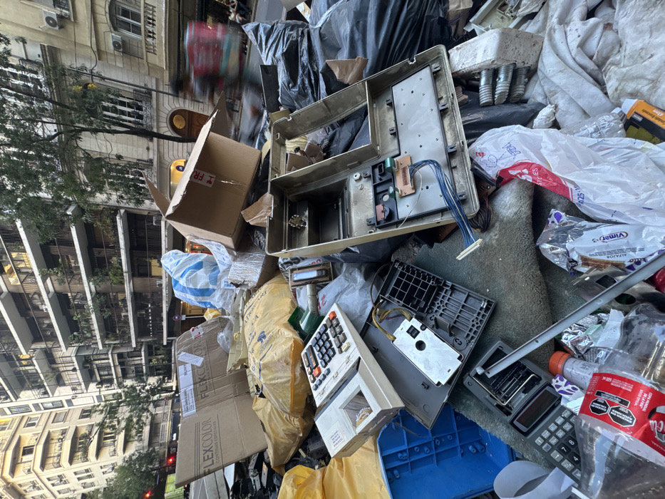 A large, dense accumulation of mixed refuse, dominated by electronic waste and packaging, occupies a street-level area in Buenos Aires, Argentina. The foreground prominently features several disassembled electronic devices, appearing as old cash registers or large calculators, with internal circuit boards, wiring, and keypads exposed. Interspersed are numerous cardboard boxes, some marked "FLEXCOLOR CORTINAS ESPECIALES" and one "FRAGILE," alongside various plastic items including yellow and black garbage bags, a blue crate, and clear plastic bottles with labels. White plastic bags, some containing "VIMET" branded packaging, contribute to the debris. The background reveals a typical Buenos Aires streetscape: a modern multi-story building on the left, and an older, ornate architectural structure with balconies and foliage on the right. A person on a motorcycle, motion-blurred, travels from right to left in the mid-ground, visibly carrying a delivery bag. Distant pedestrians are discernible on the sidewalk, and a red traffic light is visible on the far left.