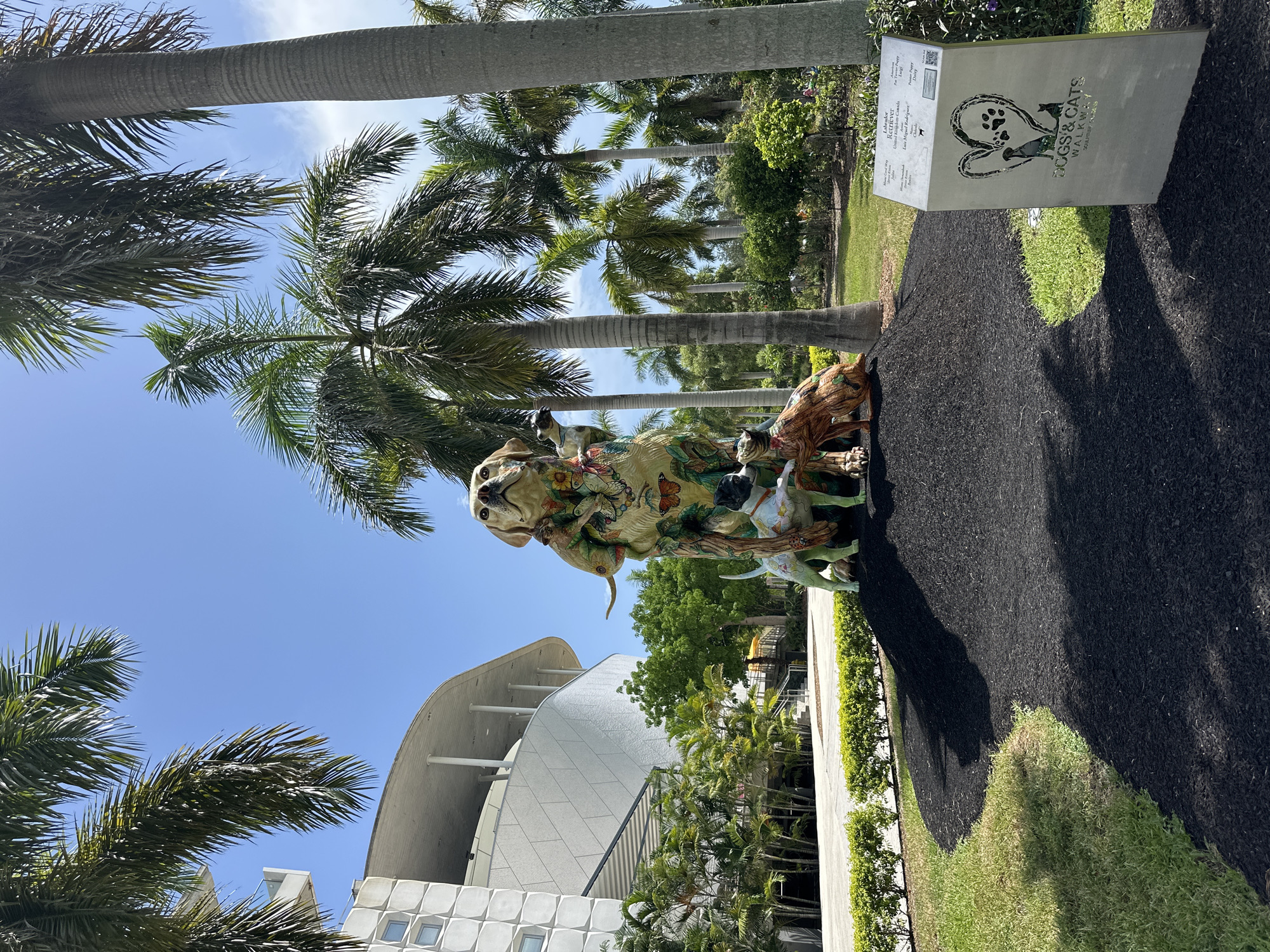 The image displays a vibrant outdoor sculpture of dogs and cats, set against a backdrop of lush greenery and palm trees, under a clear blue sky. The sculpture, situated in a park-like setting, features a large central dog figure adorned with colorful painted patterns of butterflies and flowers. Smaller dog figures are positioned around the base of the larger one. To the right of the sculpture, a sign reads "DOGS & CATS WALKWAY SCULPTURE PARK," indicating the location. In the background, a modern building with a curved roof and white facade is visible, surrounded by manicured hedges and tropical foliage. The ground around the sculpture is covered in dark mulch, transitioning to a grassy area. This scene is likely in Miami, Florida, given the prevalent palm trees and architectural style.