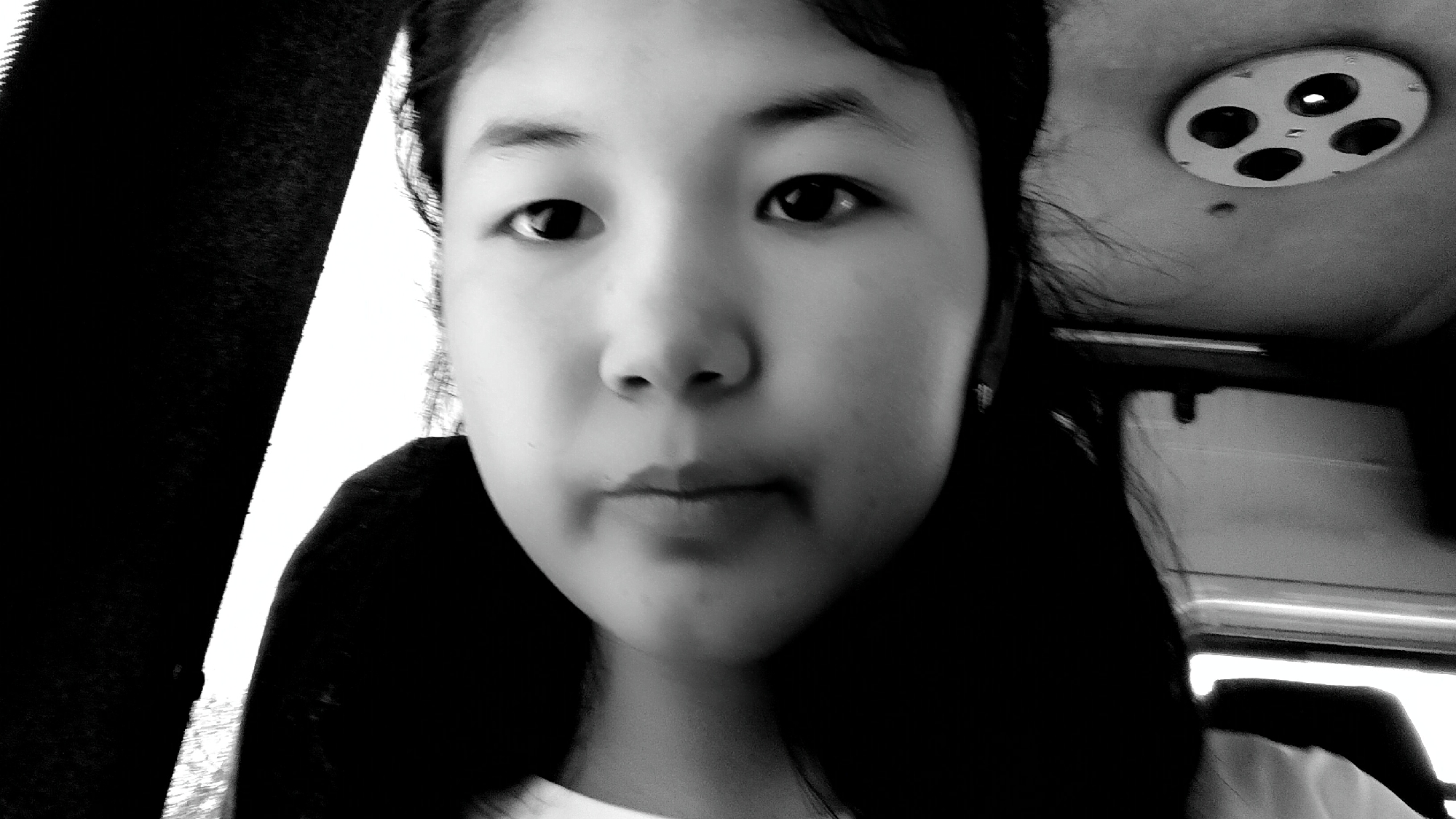A black and white close-up selfie captures a young person's face, positioned slightly off-center. The individual's gaze is directed towards the camera, with their eyebrows slightly raised. Their lips are closed, and their nose is centrally located on their face. The background is dark, with a bright light source visible on the upper right side of the image. A circular object with four dark circles is mounted on what appears to be a ceiling. The person's hair is dark and styled in pigtails that frame their face. The lighting creates harsh shadows, particularly on the left side of the face. The image is grainy, indicating a low-resolution capture or low light conditions. The overall impression is of an informal snapshot taken from a personal device. There is no indication of Kaarman, Kyrgyzstan.