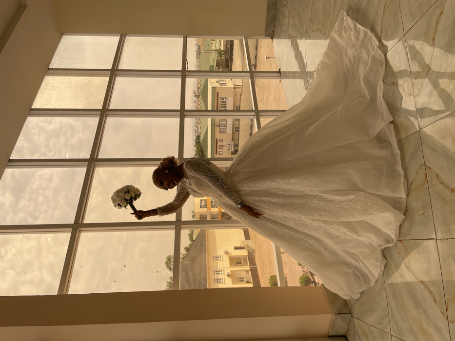 An indoor photograph depicts a female subject in a wedding gown, captured in a dynamic pose against a large, multi-pane window. The scene is located in a building in Barakin Lerawi, Nigeria.
The subject is a dark-skinned female, appearing to be a young adult, with her hair styled upwards. She is positioned towards the center-right of the frame, facing slightly to her left (viewer's left), with an open-mouthed smile. Her right arm is raised high above her head, holding a white floral bouquet composed of approximately 10-12 white flowers, possibly roses, with a dark-wrapped stem. Her left arm is slightly bent, and a silver-toned wristwatch is visible on her left wrist. She is wearing a floor-length, long-sleeved white or off-white wedding gown featuring a voluminous skirt with a considerable train, adorned with intricate beading or glitter on the bodice and skirt.
The interior environment features light-colored walls and a reflective tiled floor with a white and brown marble pattern. A light-colored pillar is visible on the far left.
The window, which spans the height of the wall and is segmented by dark frames into multiple rectangular panes, offers a view of the exterior. The sky outside appears hazy or overcast with a warm, yellowish-orange tint. Below the sky, an urban or semi-urban landscape is visible, featuring several two-story buildings. On the left, a light-colored building with a dark, sloped roof is prominent. Towards the right, other structures with light green and beige roofs are present, along with sparse vegetation. Distant hills or low mountains are faintly visible on the horizon line.