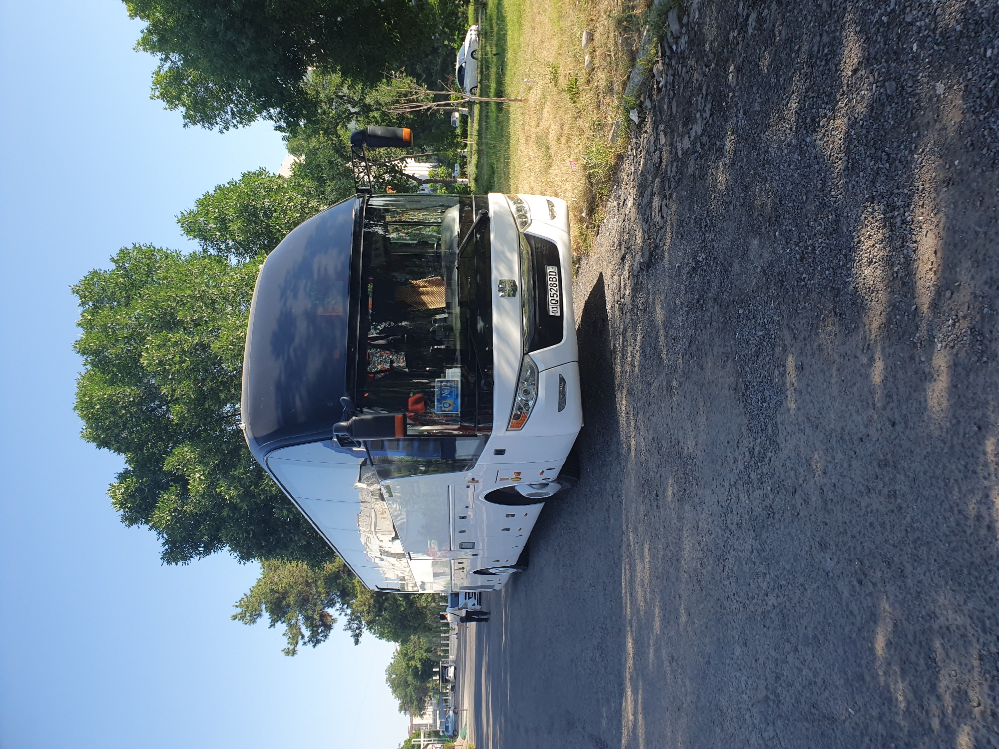A white and black tour bus with license plate "01Q528BD" is parked on a cracked asphalt road. The bus is the primary subject, positioned in the foreground and midground, taking up most of the frame. It is a modern tour bus with tinted windows and an orange turn signal on the side mirror. In the background, several green trees with lush foliage are visible, suggesting a park or a tree-lined street. The sky is clear and blue. To the left of the bus, a man in a white shirt and dark pants is walking away from the camera. Further down the road, another white car is parked. To the right of the bus, a patch of dry grass and a young tree are visible. The overall scene appears to be a sunny day in a city environment, possibly in Tashkent, Uzbekistan, given the license plate format.