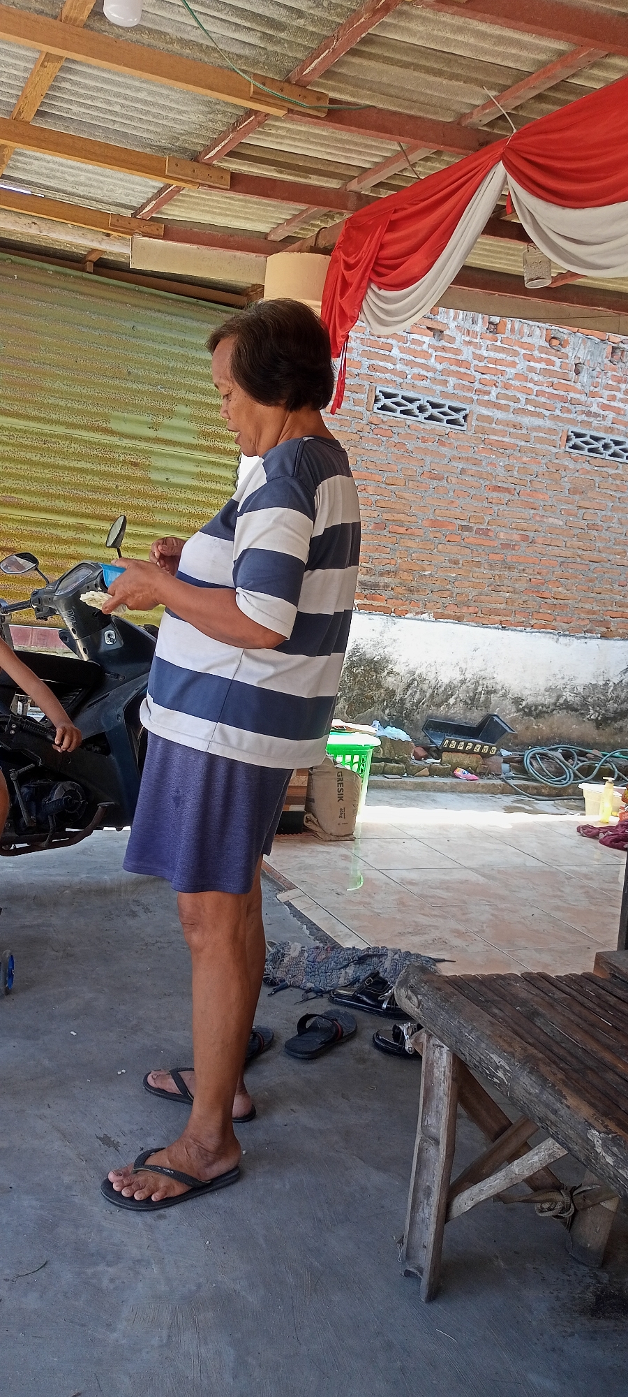 An Indonesian woman stands in an outdoor setting, likely a yard or open area, with a motorcycle beside her. She is wearing a blue and white striped t-shirt and blue shorts, with black flip-flops on her feet. She holds a light-colored object, possibly food, in her hands and appears to be in the process of eating or preparing it. Behind her, a brick wall is visible, and a corrugated metal structure forms part of the background, suggesting a rustic or unfinished building. Red and white drapery hangs from a wooden roof structure above, which is supported by exposed beams. The ground is paved with light-colored tiles. Scattered on the ground are several pairs of flip-flops and a rustic wooden bench. The scene appears to be set in Gempolklutuk, Indonesia, a location characterized by its local architecture and outdoor living spaces.