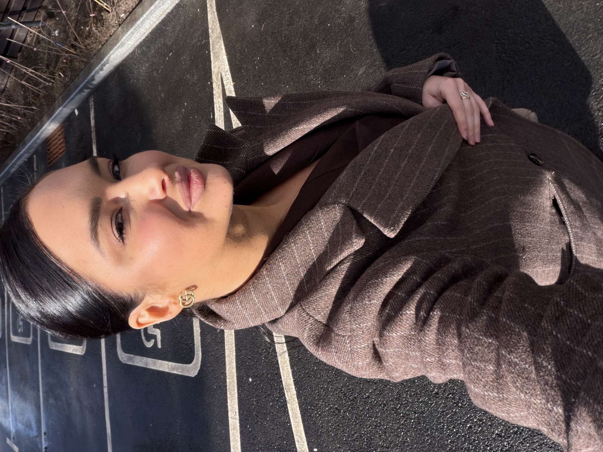 The image is a portrait of a woman dressed in a brown jacket. She is standing outdoors in Zhukovka, Russia. The ground is dark gray.