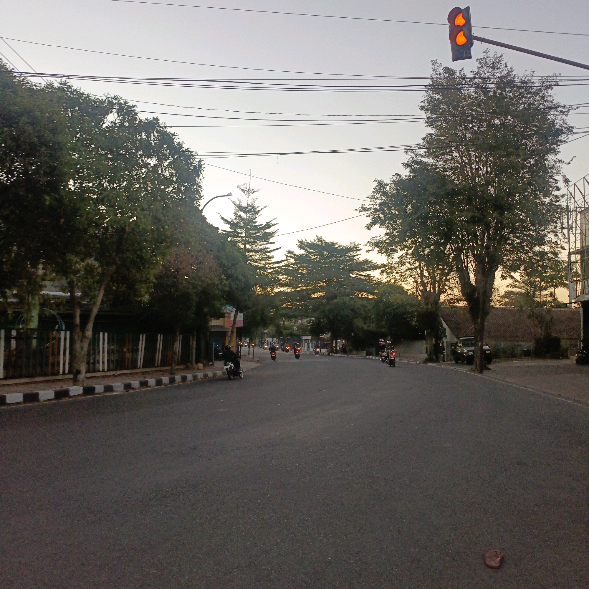The image depicts a street scene in Salatiga, Indonesia, during what appears to be either sunrise or sunset, given the soft lighting and the orange traffic light. The road is asphalt, with a distinct curb on the left side featuring alternating black and white painted sections. Several trees line the left side of the road, casting shadows. On the right side, more trees are visible, and a building with a brown tiled roof is partially obscured. The sky is a pale gray, with visible power lines stretched across it.
In the foreground and mid-ground, several motorbikes are in motion or parked. On the left, a single motorbike is stopped near the curb, ridden by a person wearing a dark helmet and jacket. Further down the road, more motorbikes are scattered, with riders visible on some. The traffic light at the intersection ahead is illuminated with a solid orange light. A few more vehicles, including what appears to be a pickup truck and another car, are parked on the right side of the road. A single red flip-flop lies on the asphalt in the lower right corner of the image. The overall impression is of a typical urban street with moderate traffic activity.