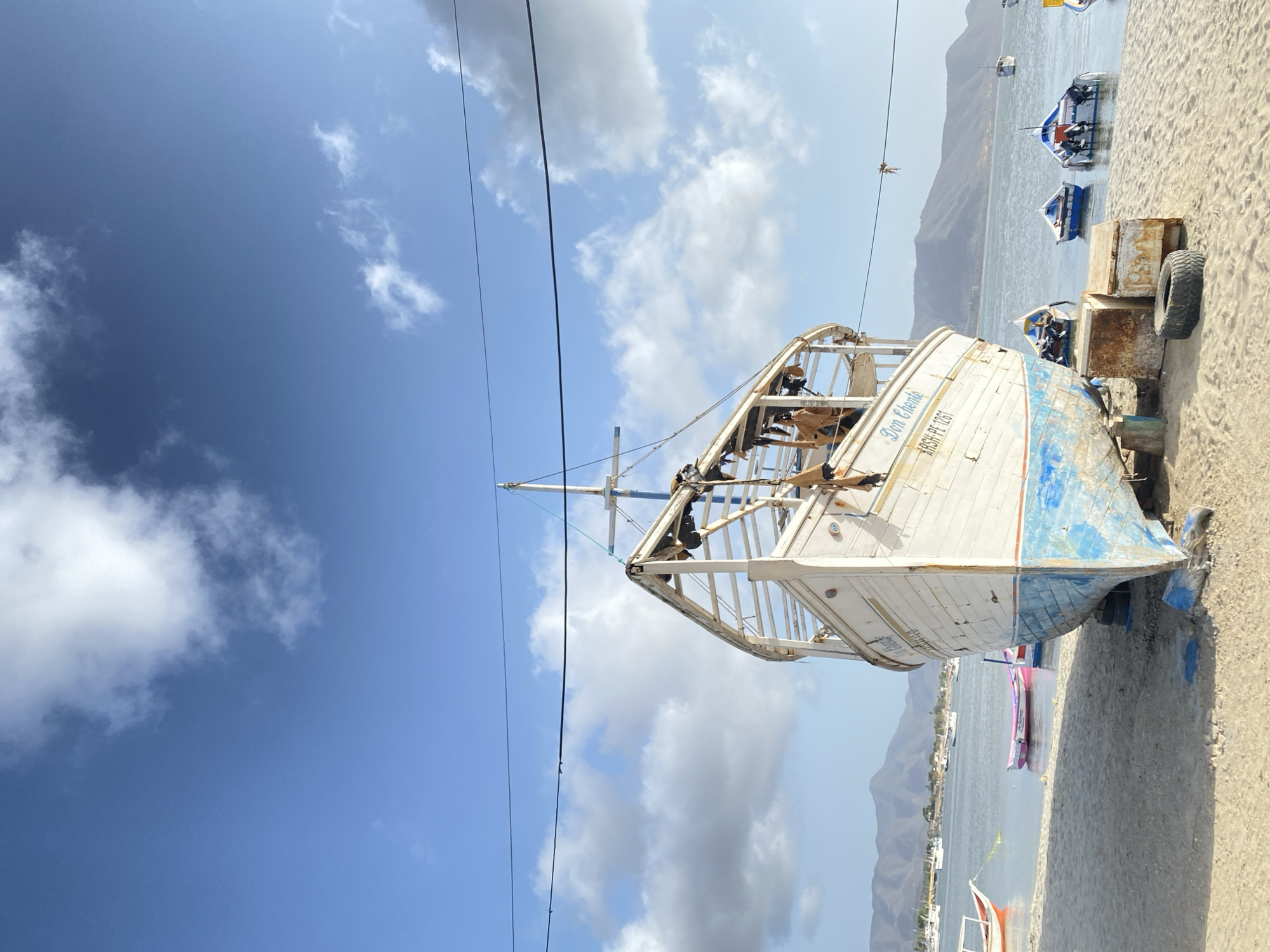 An old, weathered boat, likely a fishing vessel, rests on a sandy beach in Juan Griego, Venezuela. The boat's hull is predominantly white with patches of faded blue paint. "Don Chente" is written on the side of the boat. A makeshift wooden structure extends from the boat. It's likely undergoing repairs or has been abandoned. The beach is visible with scattered smaller boats at the edge of the water. In the background are rolling hills. The sky above is blue with scattered clouds. Power lines run across the top part of the image.
