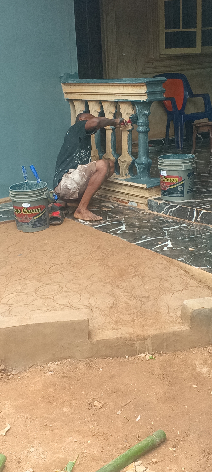 The image depicts an outdoor scene in Eziudo, Nigeria, where an adult male is actively painting a balustrade. The setting is likely the porch or veranda of a residential building.
The man, of dark complexion, is dressed in a dark t-shirt and shorts, both visibly splattered with paint, indicating his current work. He is barefoot and crouched low, diligently applying a dark teal or blue-green paint to the decorative spindles of the balustrade using a small brush. His slippers are placed on the tiled floor beside him, next to a grey paint bucket.
The balustrade itself is made of a light-colored material, possibly concrete, with a contrasting beige or light yellow top rail. The house wall in the background is painted a similar teal or blue-green, suggesting a coordinated color scheme. Two grey paint buckets are prominent in the foreground. One bucket, closer to the man, contains paint brushes with blue handles. Both buckets display "Mega Crown Paints" branding, and visible text also includes "Emulsion Paints," "ICI Paints," and "30 kg."
The floor of the porch is covered in dark, possibly black or dark grey, tiles with prominent white veining. This tiled area transitions to a light brown pathway, which features an elaborate, embossed pattern of interconnected circles. Beyond this patterned pathway, the ground consists of loose, reddish-brown soil, with a green stick, possibly bamboo, lying in the immediate foreground. In the background, past the balustrade, a section of the house wall, a window, and an orange and blue plastic chair are visible. The scene is well-lit, indicating it is daytime, and the man appears focused on his task.