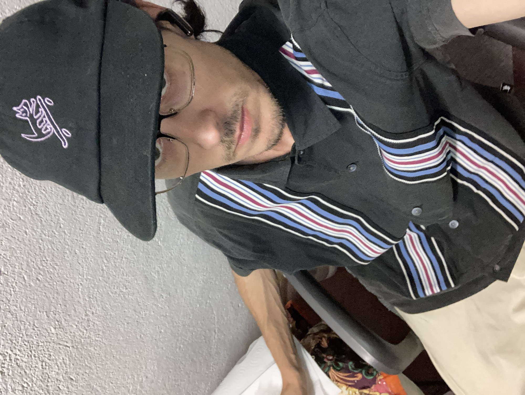 A medium shot captures a person wearing a black baseball cap with a pink embroidered cat riding a skateboard and glasses. They are seated in a chair, seen from a slightly elevated angle, facing forward. The person has a mustache and a goatee. They are wearing a black collared shirt with vertical stripes in white, blue, and maroon on the front and sleeves. Khaki-colored pants are visible below the shirt. The background consists of a textured, white wall and a glimpse of a colorful fabric object on the left. The scene appears to be indoors, potentially in a residential or office setting. The image quality is slightly grainy.