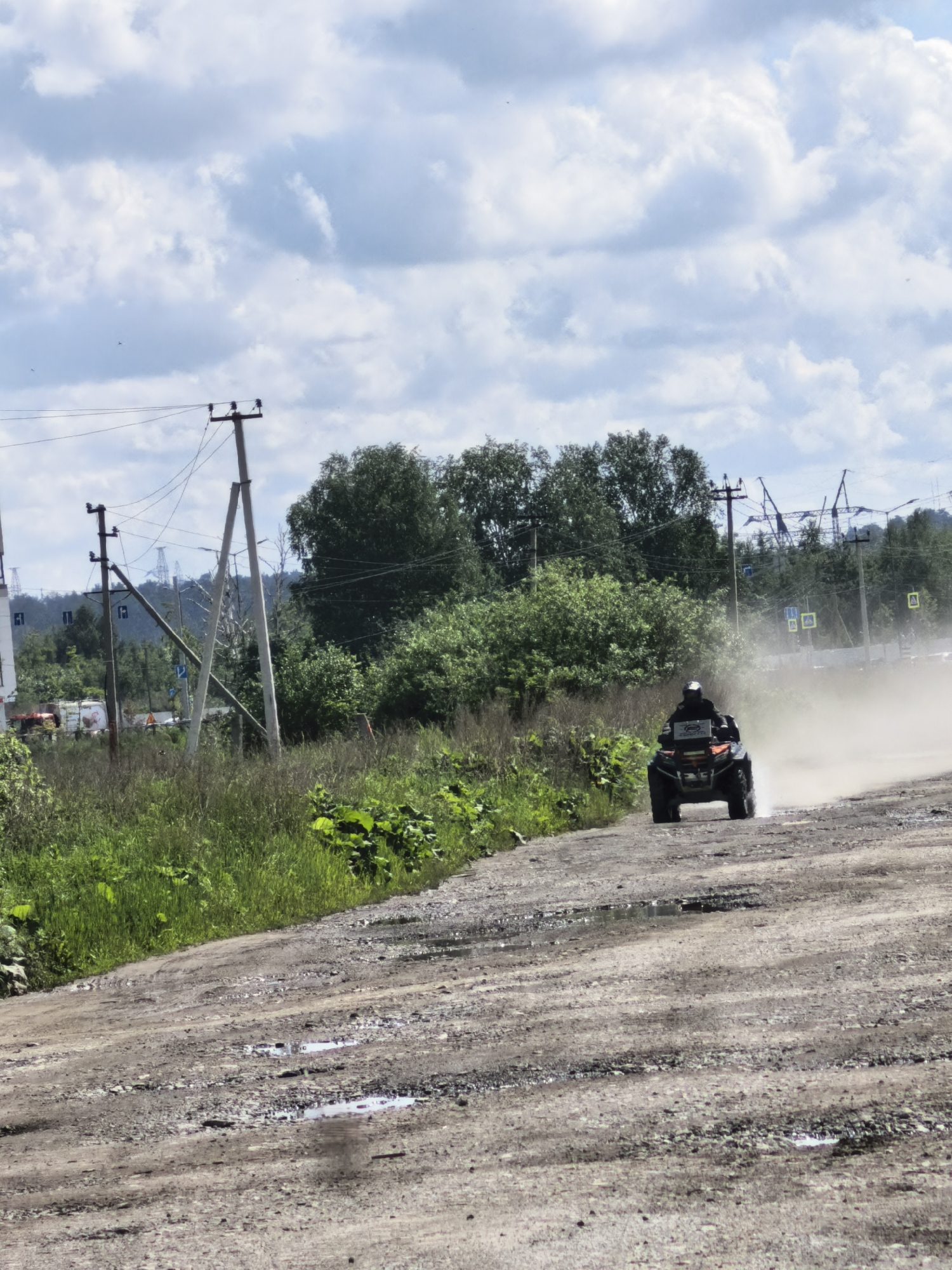 An ATV is seen kicking up a cloud of dust as it rides on a dirt road. The rider is wearing a helmet. The road is unpaved and has several puddles. On the left side of the road, there is tall green grass and trees. In the background, there are power lines, more trees, and some industrial structures. The sky is cloudy.
