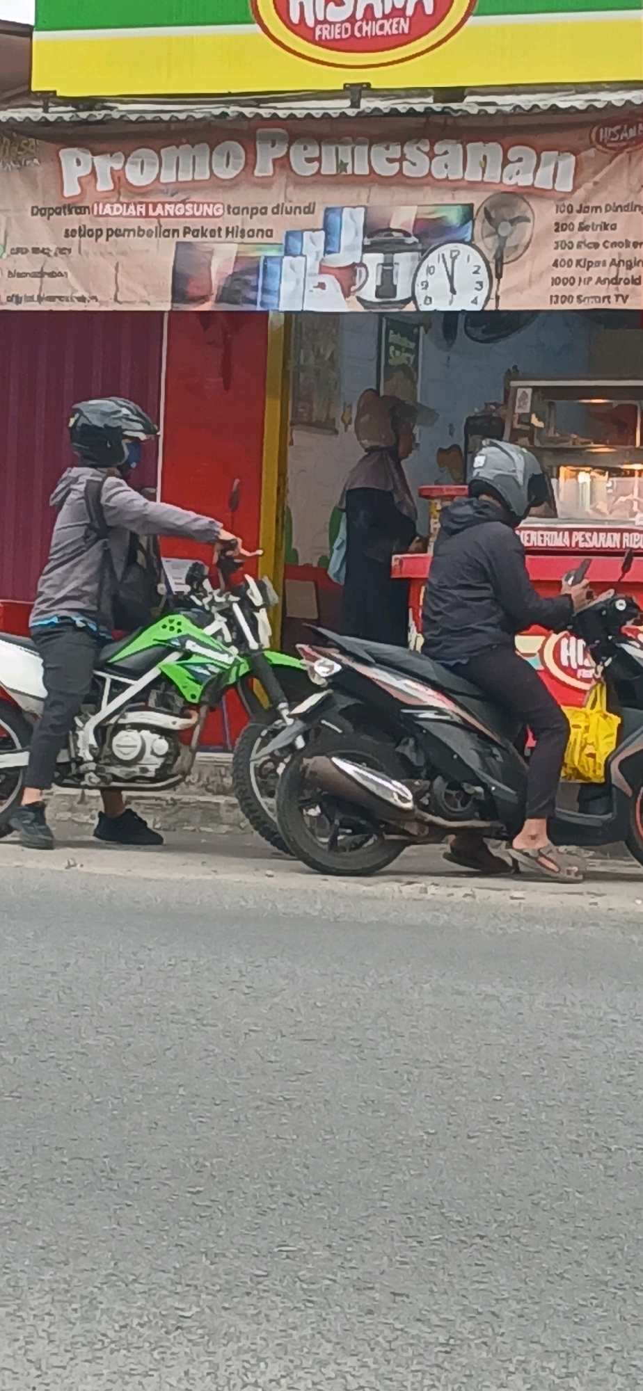 The image depicts a street scene in Cirebon, Indonesia, focusing on a banner advertising a promotion. The banner, positioned above a storefront, announces "Promo Pemesanan" (Order Promotion) and details prizes for purchasing "Paket Hisana" (Hisana Package). Prizes listed include a 100 Jam Dinding (100 Wall Clock), 200 Botolika (200 Bottles), 300 Rice Cooker, 400 Kipas Angin (400 Fans), 1000 HP Android (1000 Android Phones), and 1300 Smart TV.
In the foreground, two individuals are seated on motorcycles. The person on the left, wearing a grey hoodie, black pants, and a helmet, is positioned on a green and white dirt bike. The person on the right, also wearing a helmet and dark clothing, is on a black scooter, holding a smartphone. Behind them, a person in a hijab is visible near the storefront, possibly interacting with a customer or employee. The environment appears to be an urban setting with buildings and a paved road. The banner is illuminated by natural light, suggesting it's daytime.