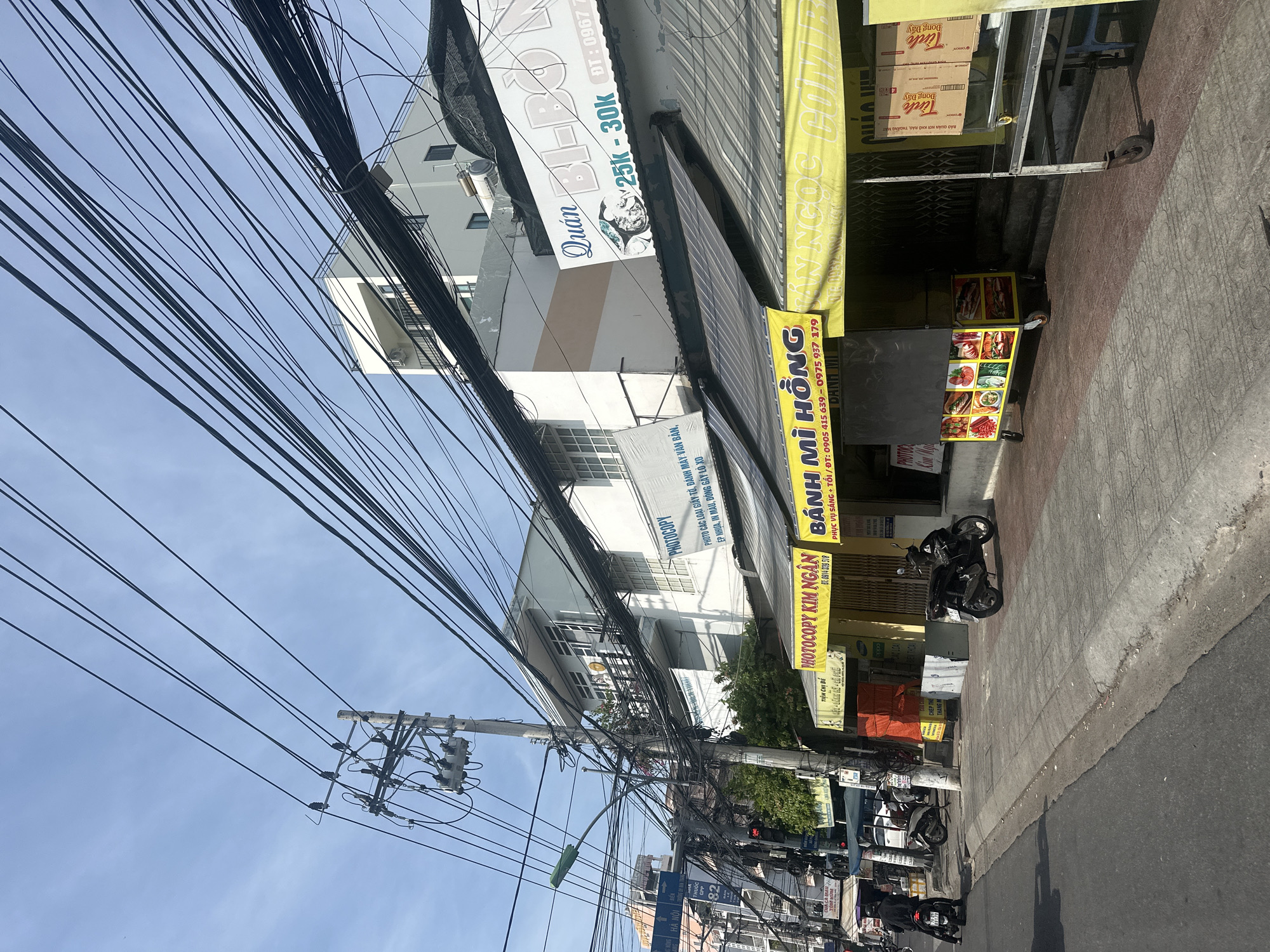 The image depicts a street scene in Bac Nha Trang Ward, Vietnam. A utility pole with numerous electrical wires is prominent on the left side of the frame, with a traffic light visible below it. Several businesses line the sidewalk, including a "PHOTOCOPY KIM NGAN" and a food stall with a yellow awning labeled "BANH MI HONG." Another business with a large sign featuring a fish and the text "Quan BI-BO Ngu" is also visible. Two motorcycles are parked on the sidewalk, and one is being ridden on the street. The buildings are multi-story, and the sky is clear and blue. The overall scene is characteristic of a bustling urban environment in Vietnam.
