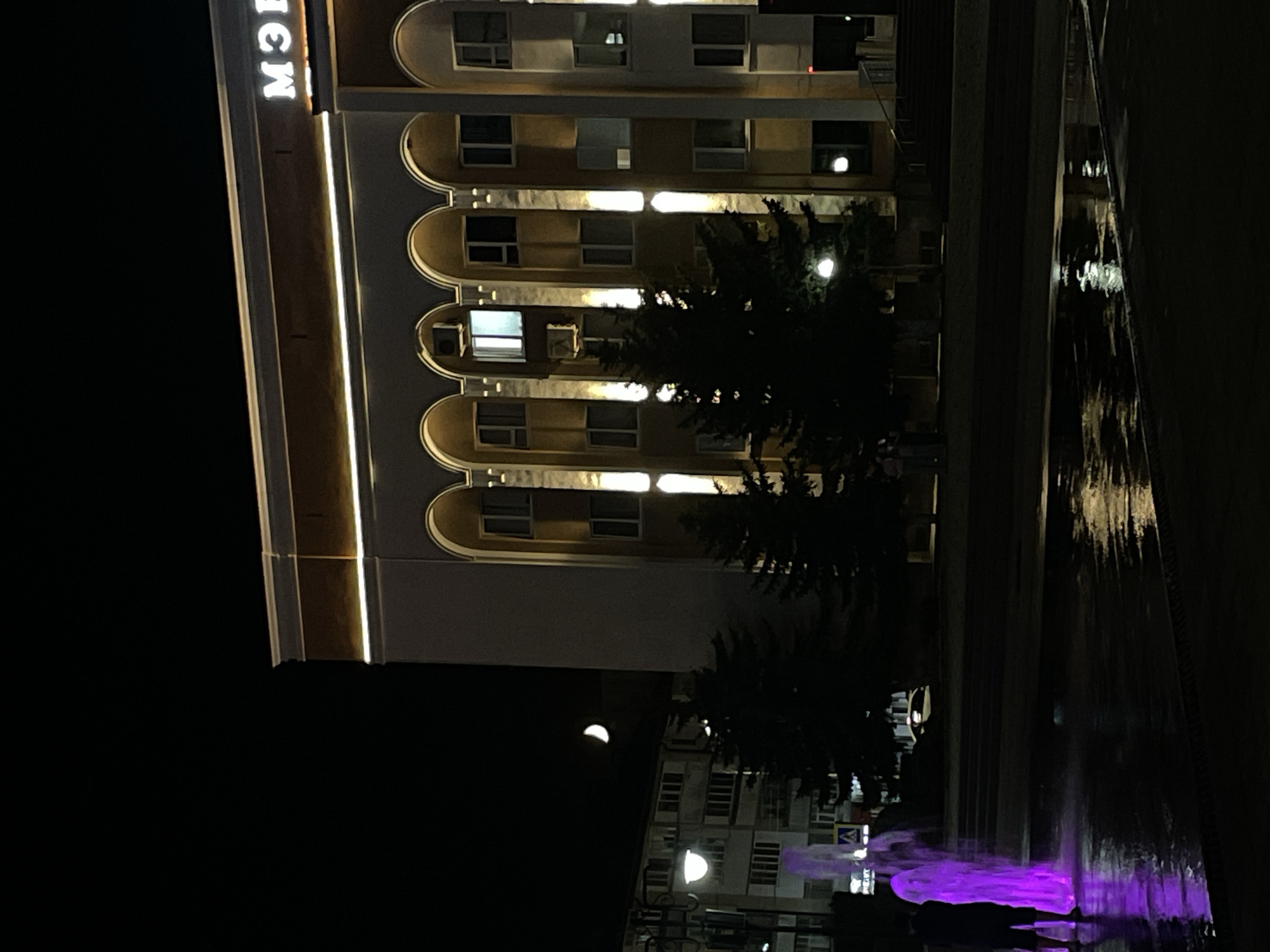 At night, in Cherkessk, Russia, a broad, dark street is illuminated by a bright, purple fountain in the foreground, reflecting on the wet pavement. Two silhouetted figures stand near the fountain, facing away from the camera. Behind them, a multi-story building with arched windows and illuminated vertical lighting dominates the scene. The building has a sign with white, glowing letters that read "МЭЗ" at the top. To the left of the building, a crescent moon is visible in the dark sky above a residential building. Several dark evergreen trees are interspersed between the fountain and the main building, and a street lamp is visible to the left of the frame. The overall scene depicts an urban night view with architectural lighting and a decorative water feature.