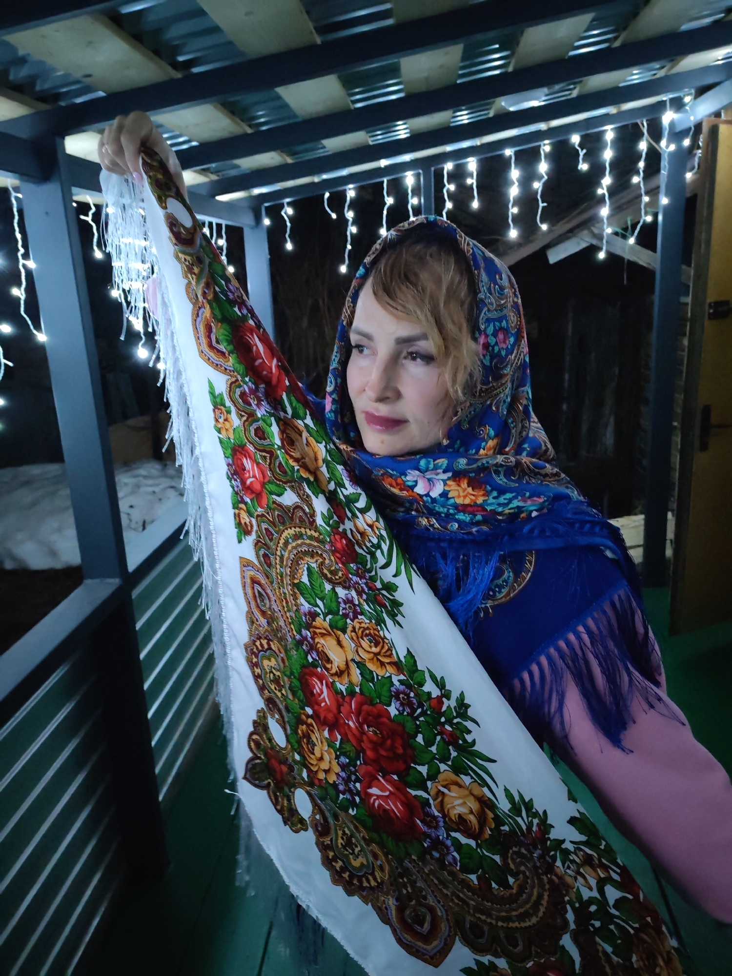 A woman stands in an outdoor structure adorned with strung lights. She is holding up a large shawl with a white background and a floral pattern primarily composed of roses in shades of red, orange, and yellow. She is also wearing a blue headscarf and a pink long-sleeved shirt. The structure has a metal roof and what appears to be a slatted wall behind her. Patches of snow can be seen in the background. There is a dark fence visible behind the snow. The location is in the city of Chaikovsky, Russia.