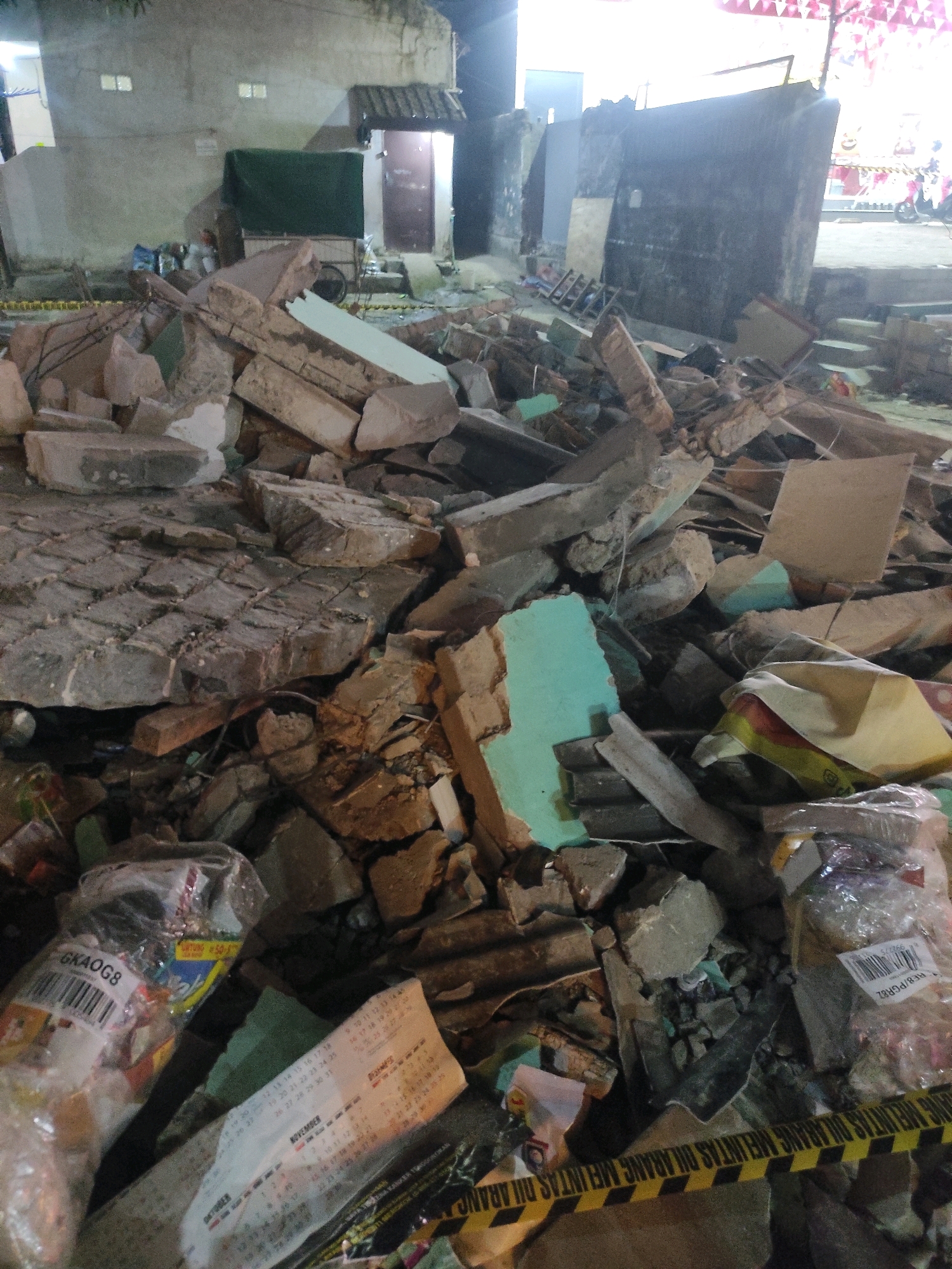 The image depicts a scene of destruction in Padalarang, Indonesia, likely a residential area, following an event that caused structural collapse. The foreground is dominated by a large pile of debris, consisting of broken concrete slabs, bricks, and other building materials. Scattered within the rubble are various objects, including plastic bags containing items, torn paper, and what appears to be caution tape with yellow and black diagonal stripes and text in Indonesian.
In the background, a building with a light-colored facade and a dark-tiled roof is visible. It has at least two small, rectangular windows. A dark green tarp covers a portion of what appears to be a cart or stall against the building. To the right of the main building, a dark-colored wall or structure stands, with some debris at its base. Further to the right, a bright, festive banner with red and white elements is visible, along with a portion of a motorcycle. The overall lighting suggests it is either dusk or nighttime, with artificial lights illuminating the scene in some areas. No people are visible in the immediate foreground or mid-ground, though movement may be implied by the presence of the motorcycle in the distance. The scene suggests a recent disaster or demolition, with the caution tape indicating an area that is unsafe or restricted.