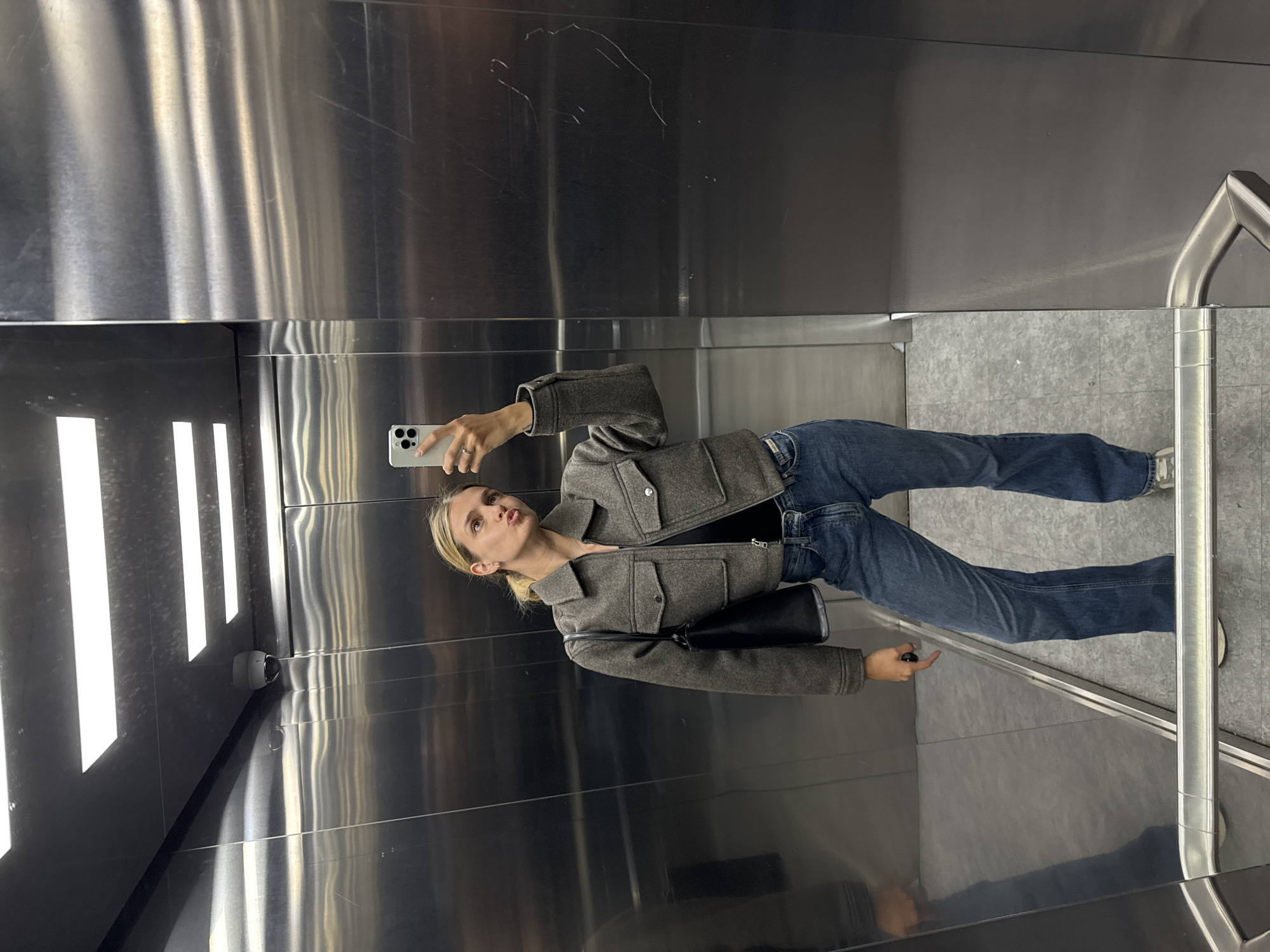 A female stands inside a modern elevator with stainless steel walls and a tiled floor, taking a selfie with her smartphone. She is wearing a gray jacket, blue jeans, and carries a black purse. Her hair is pulled back. The elevator features vertical white lighting on one side and a handrail on the other. A security camera is visible on the wall. The location appears to be in the city of Unknown, Unknown.