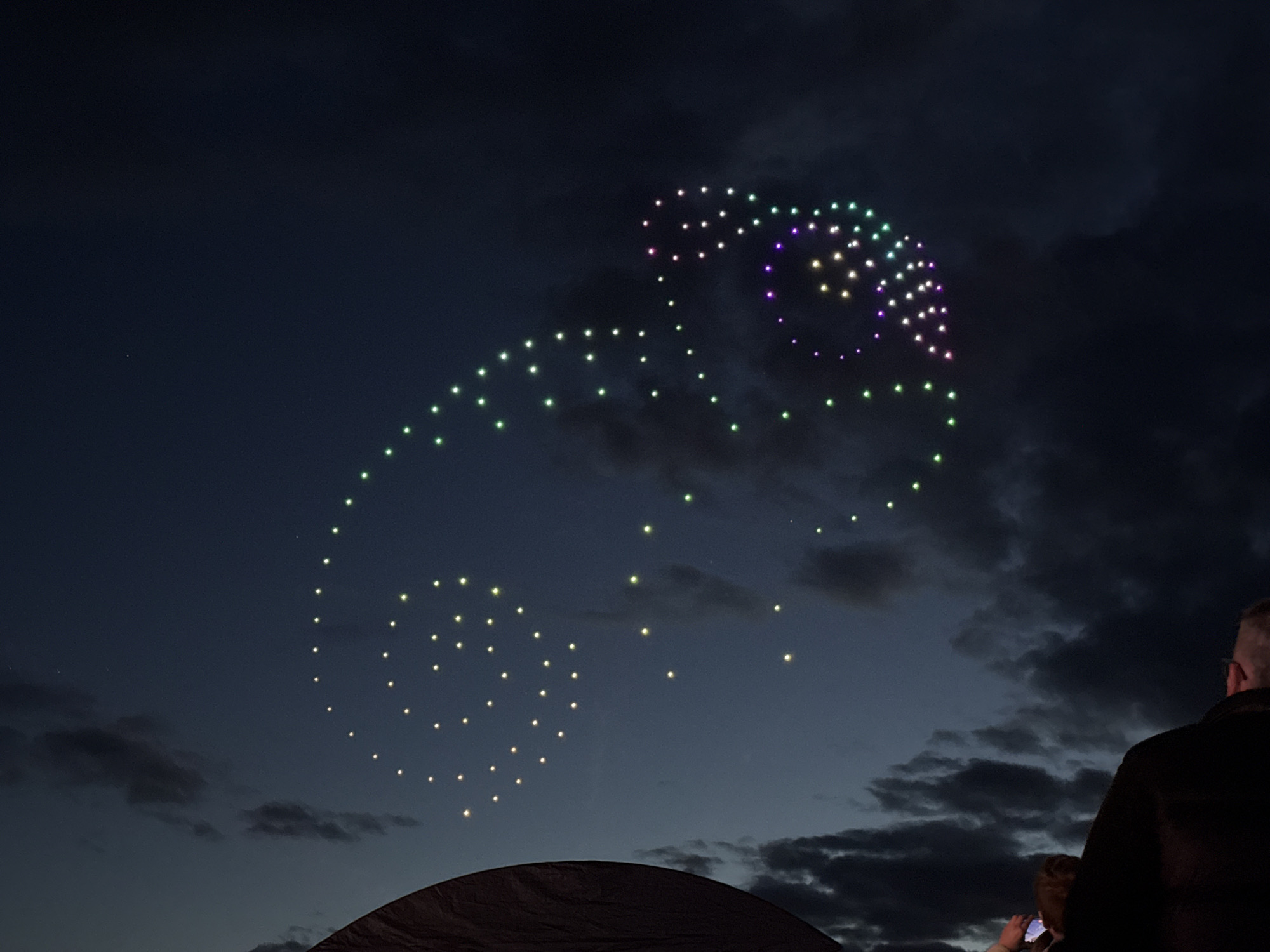 At night in South Norfolk, United Kingdom, a drone light show displays a chameleon shape in the sky. The chameleon's outline is created by numerous small lights, predominantly green and white, with purple accents. The lights are arranged to mimic the form of a chameleon, featuring a curled tail. Clouds are visible in the background. In the foreground, a dark, possibly tent-like, structure is partially visible. To the right, two individuals stand observing the spectacle. One of them appears to be holding up a phone, possibly taking a photo or video.