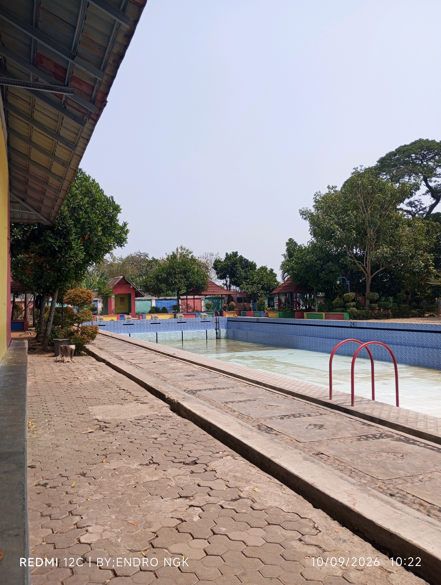 An outdoor swimming pool area is depicted on a sunny day. The pool itself is largely empty, with a shallow layer of water at the bottom. Red metal ladder rungs are visible in the pool. The pool is surrounded by a blue tiled edge and a paved walkway. To the left of the pool, a yellow building with a corrugated metal roof is partially visible. Beyond the pool, there are trees, colorful structures that appear to be small buildings or gazebos, and a fence. The ground is paved with interlocking concrete blocks. The lower right corner of the image displays text indicating "10/09/2026 10:22". The lower left corner displays "REDMI 12C | BY; ENDRO NGK".