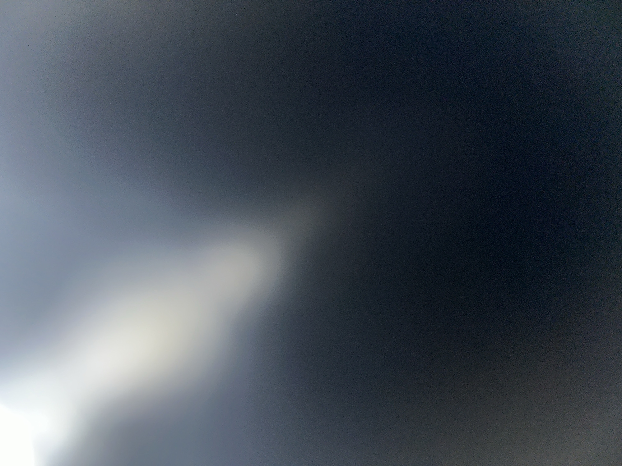 The image shows a dark, blurry scene with a bright, soft light emanating from the bottom left corner, casting a hazy glow across a portion of the frame. The light source itself is not visible. The majority of the image is a dark, gradient of grey to black, suggesting a low-light environment. There are no discernible subjects, objects, or a clear scene. The abstract nature of the image, with its focus on light and shadow, offers no specific clues about the time of day, weather, or location, other than the provided context of Belgrade, Serbia. No text is visible.